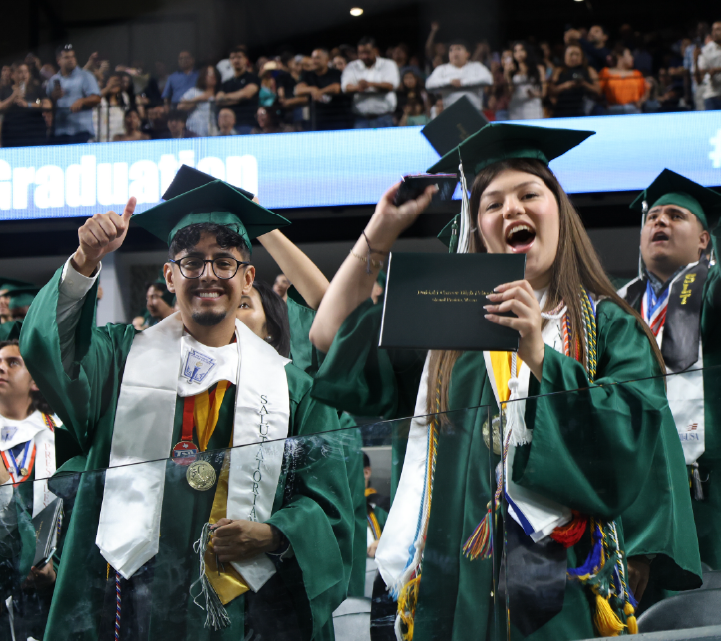 A group of graduates in green gowns and mortarboards celebrating their accomplishment, with a large audience visible in the background.