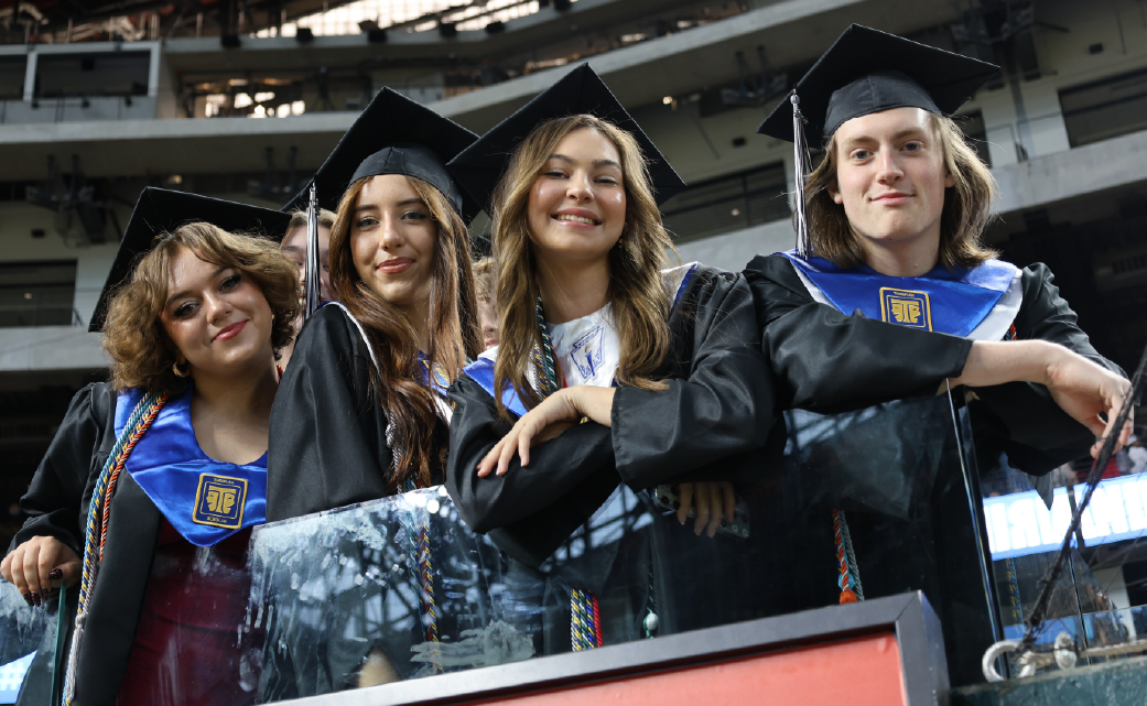 A group of four young adults in graduation gowns and caps, smiling and posing together in what appears to be a large indoor venue or auditorium.