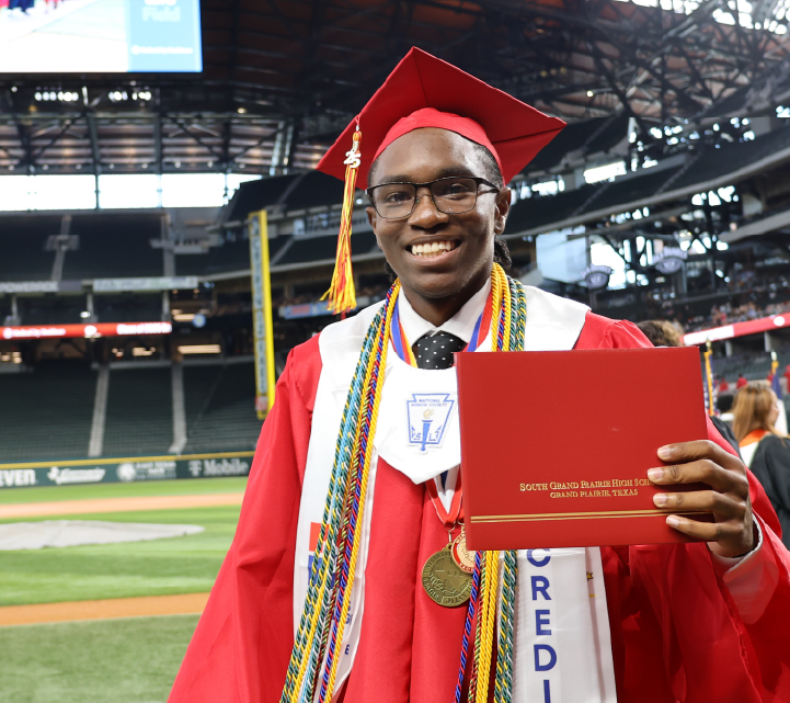 A smiling young man in a red graduation gown and cap holds a diploma while standing on a baseball field with a stadium in the background.