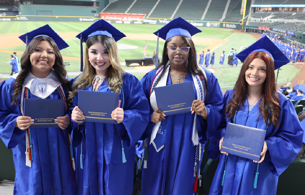 Four young women in blue graduation gowns and caps are standing together, holding their diplomas, in what appears to be a sports stadium or arena.