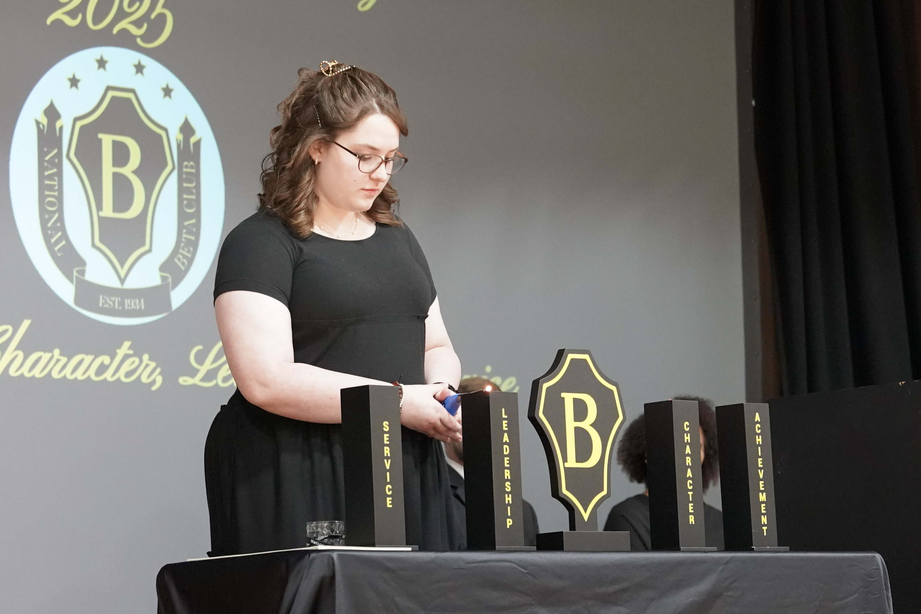 Girl on stage lights candle on table with Beta Club emblem