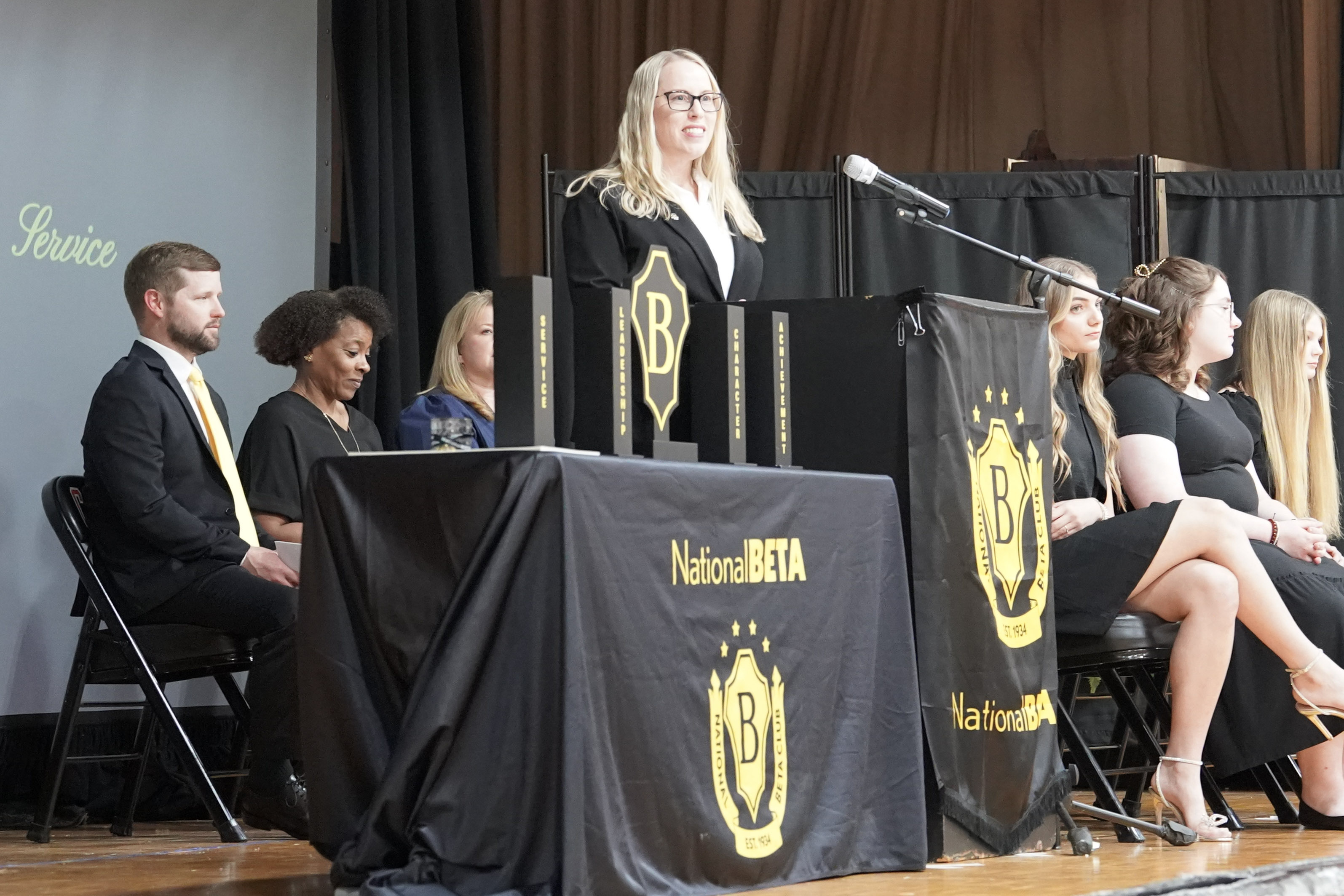 Woman in black clothing stands behind podium with Beta Club emblem with students and adults seated in background
