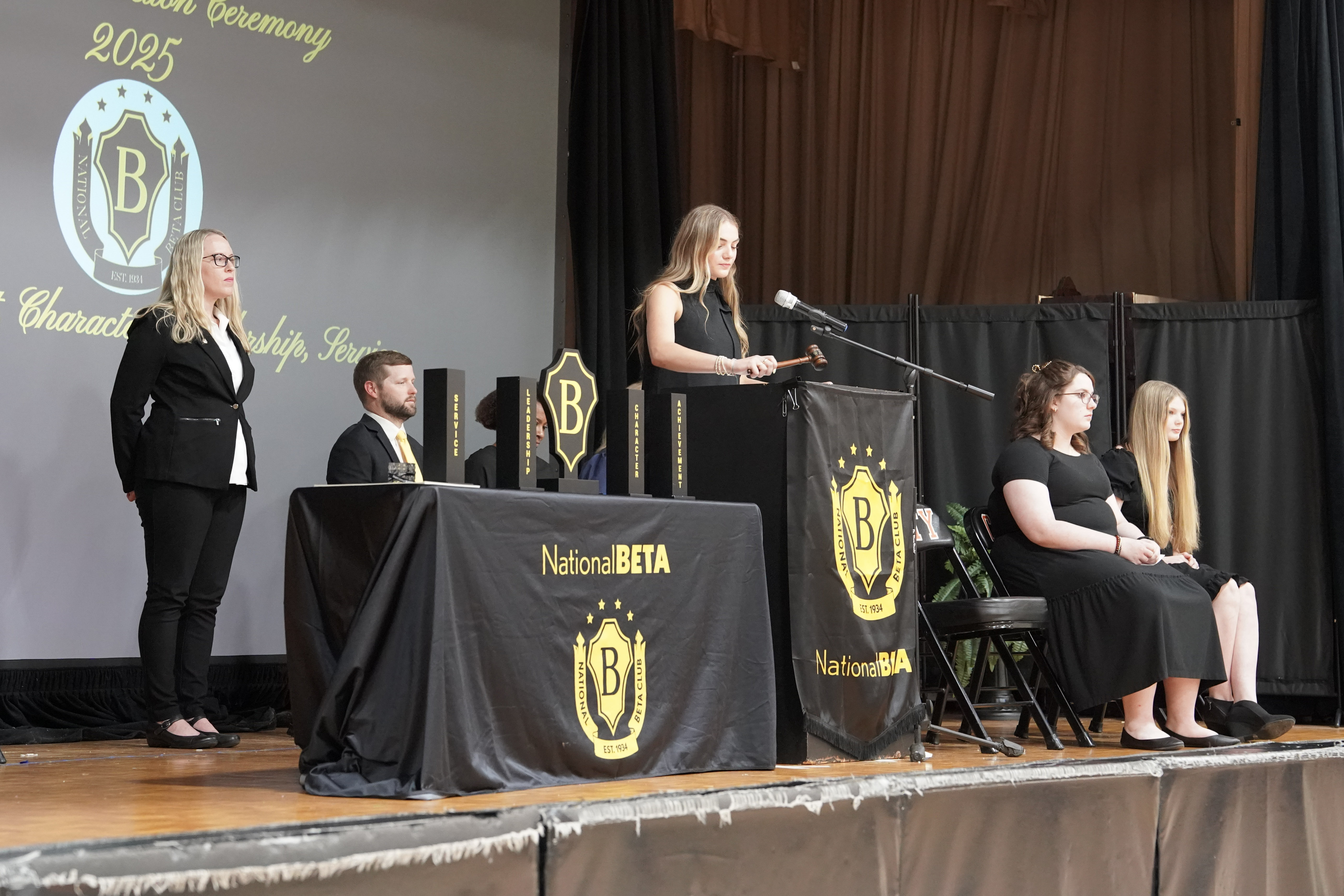 Student in black dress behind podium with other students seated and faculty members behind her