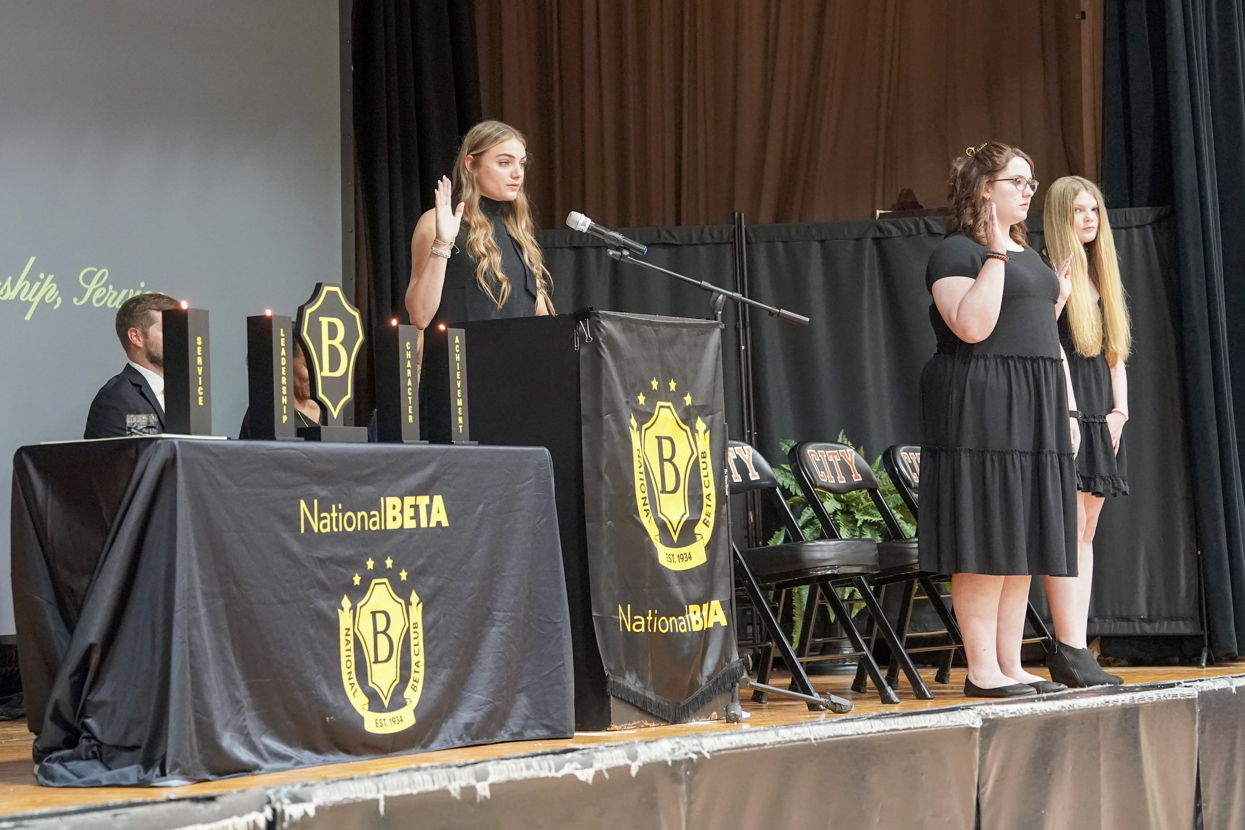 Girls in black dresses stand on stage with right hands raised and Beta emblem on podium