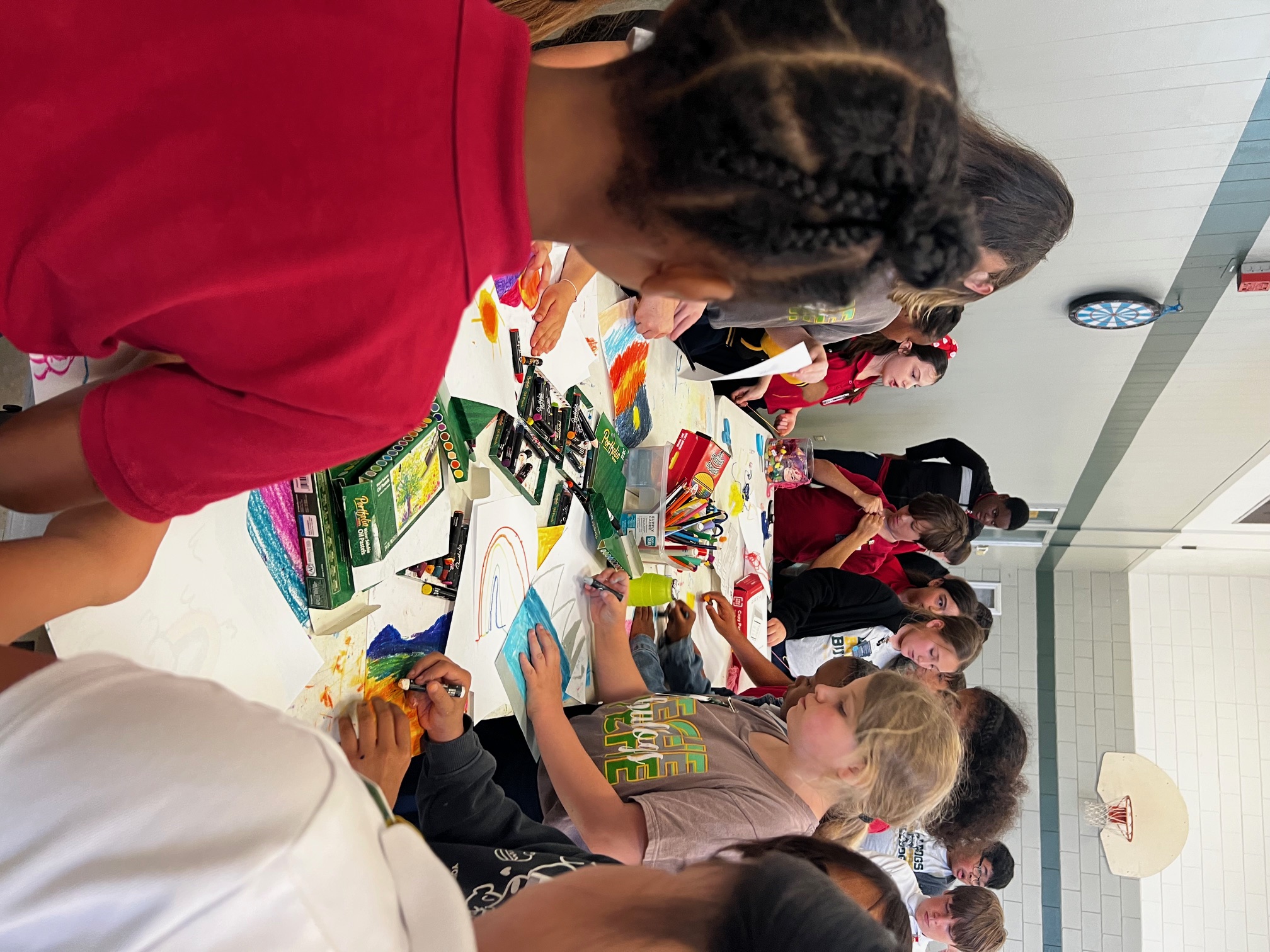 Children in a classroom are gathered around a long table covered in art supplies and colorful drawings, with a basketball hoop visible in the background.