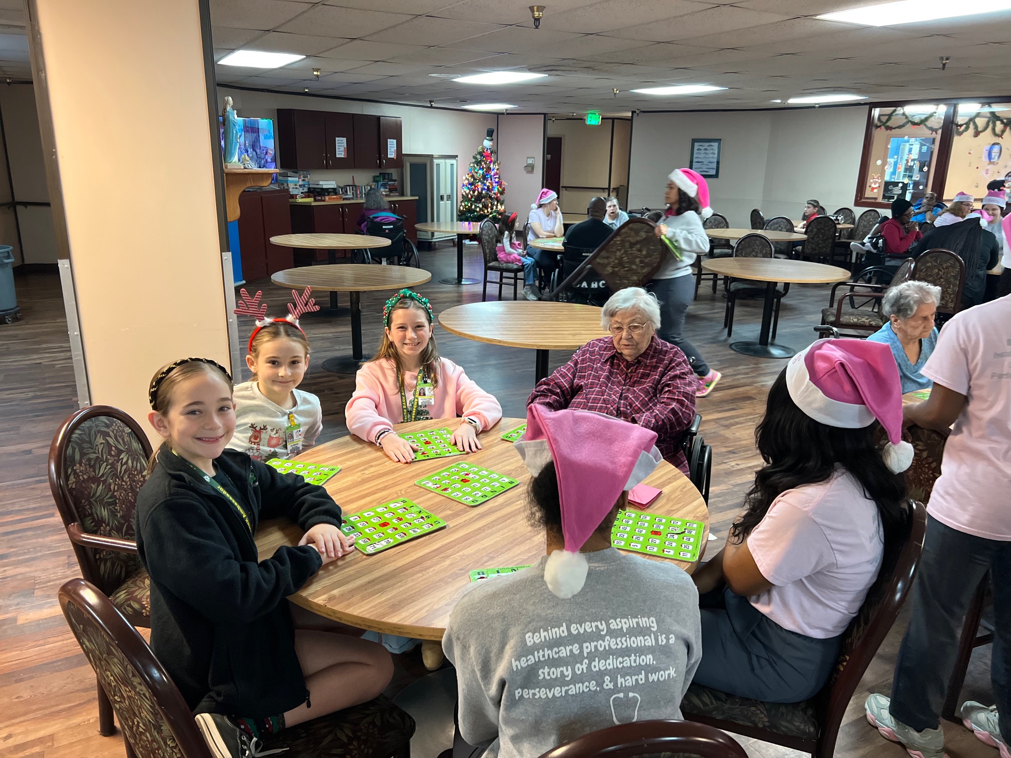 In the foreground, a group of people, some wearing Santa hats, play bingo around a round table, while in the background, a decorated Christmas tree and other residents are visible in a common room.
