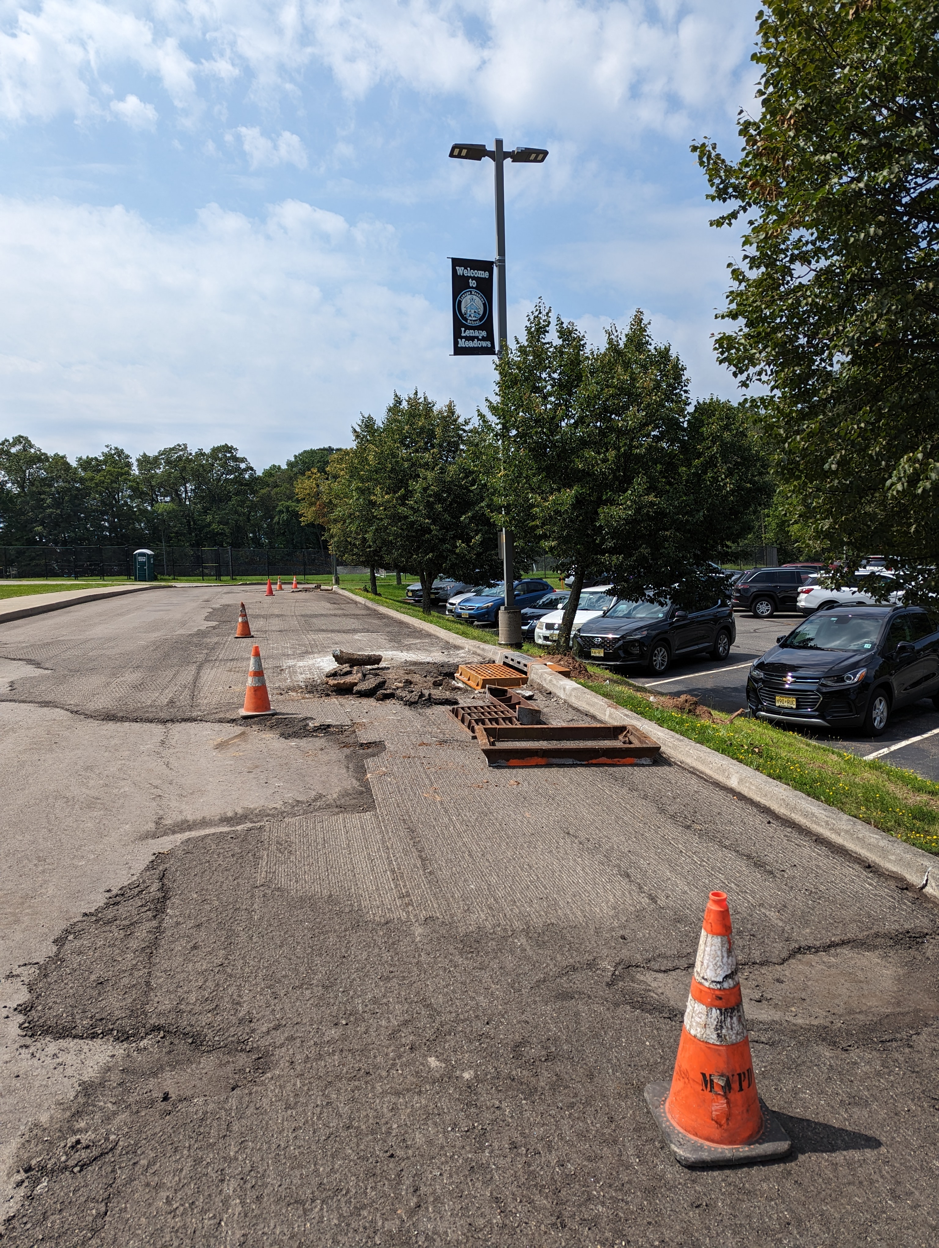 Orange traffic cones mark a patch of damaged asphalt in the foreground, while a parking lot with cars and trees stretches into the background under a bright sky.
