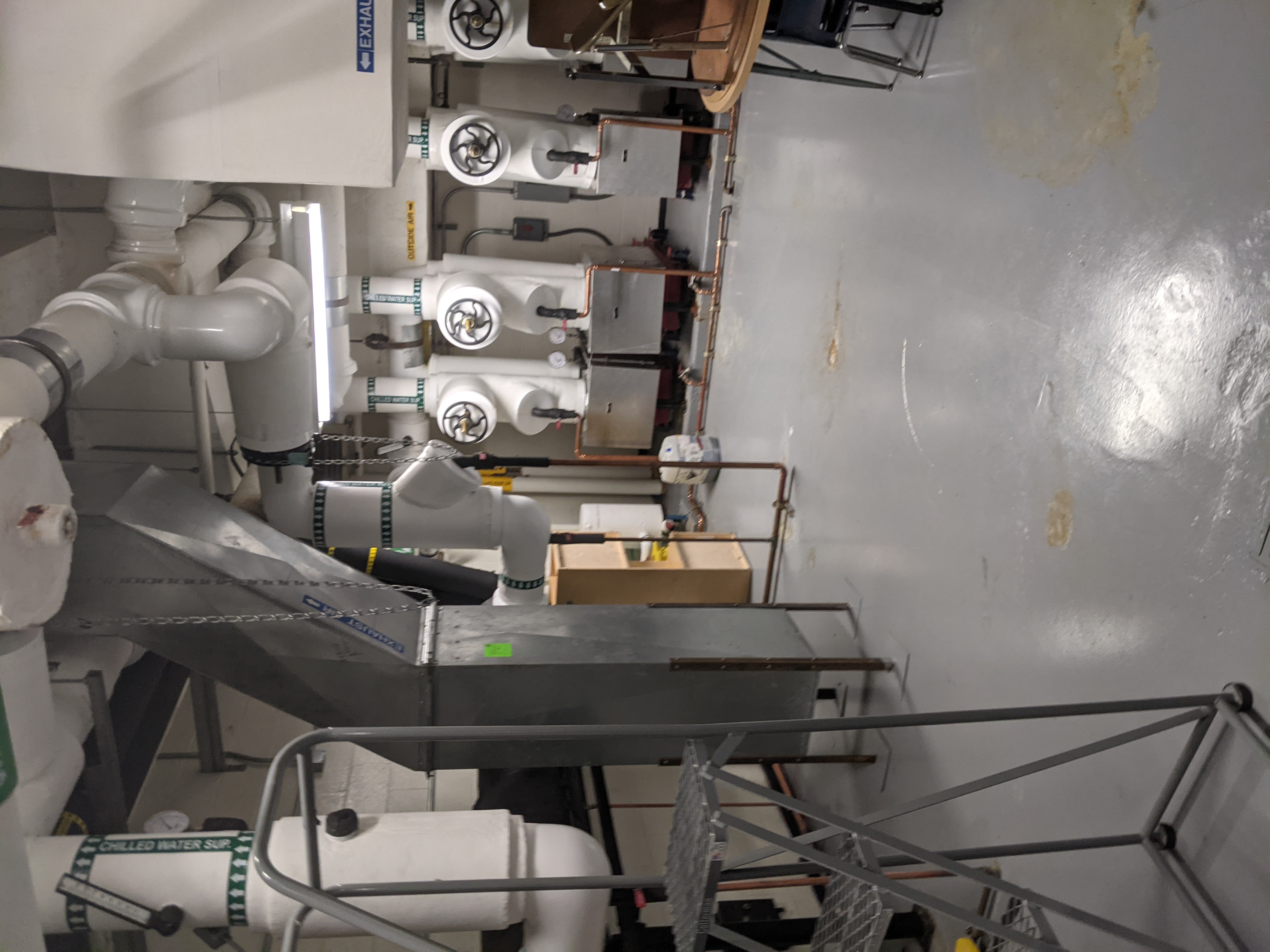 A metal ladder and ductwork are in the foreground, while large white machinery with pipes is in the background.