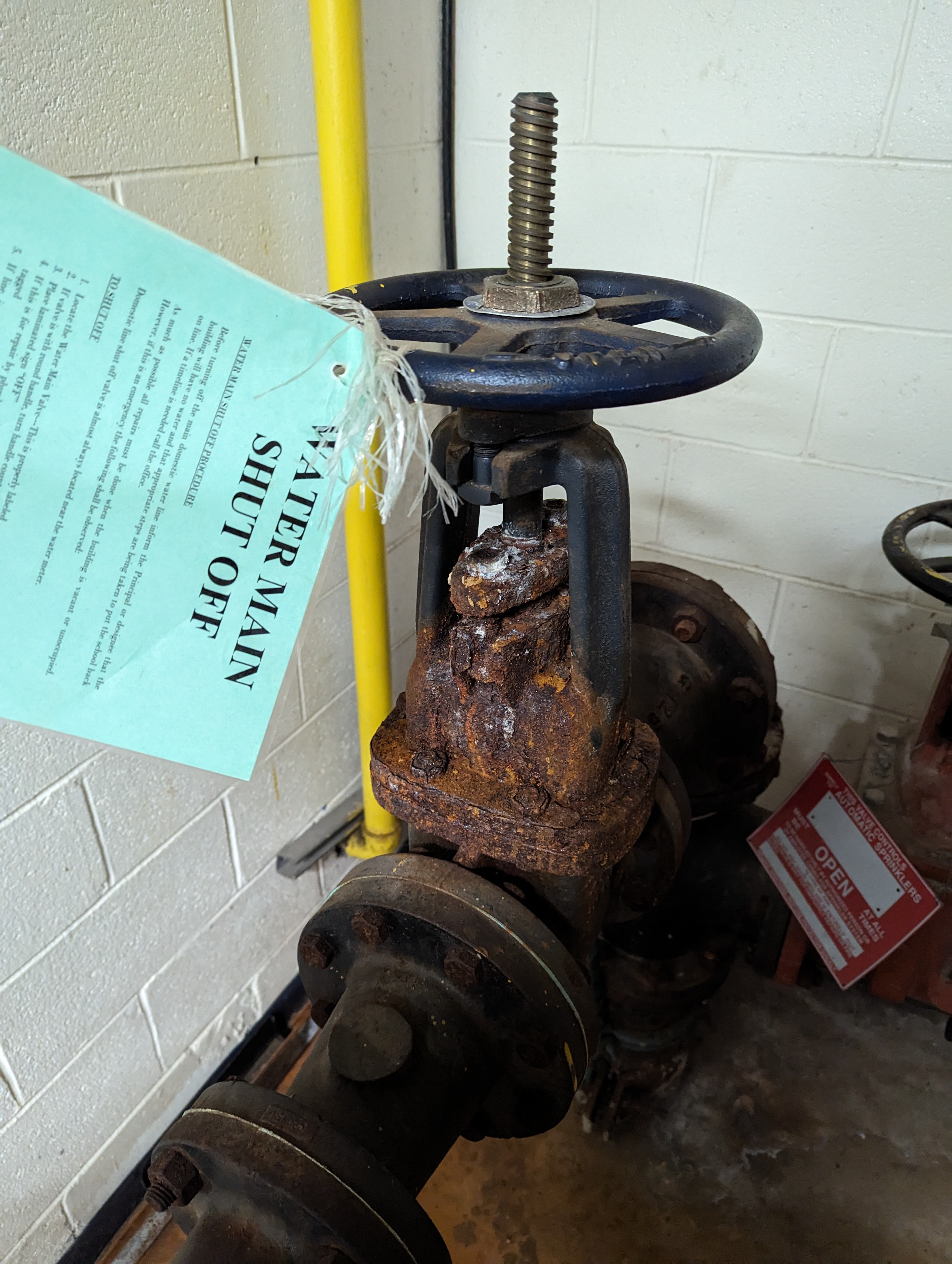 A rusty water main shut-off valve with a "WATER MAIN SHUT OFF" tag hangs in the foreground, while a yellow pipe and a brick wall form the background.
