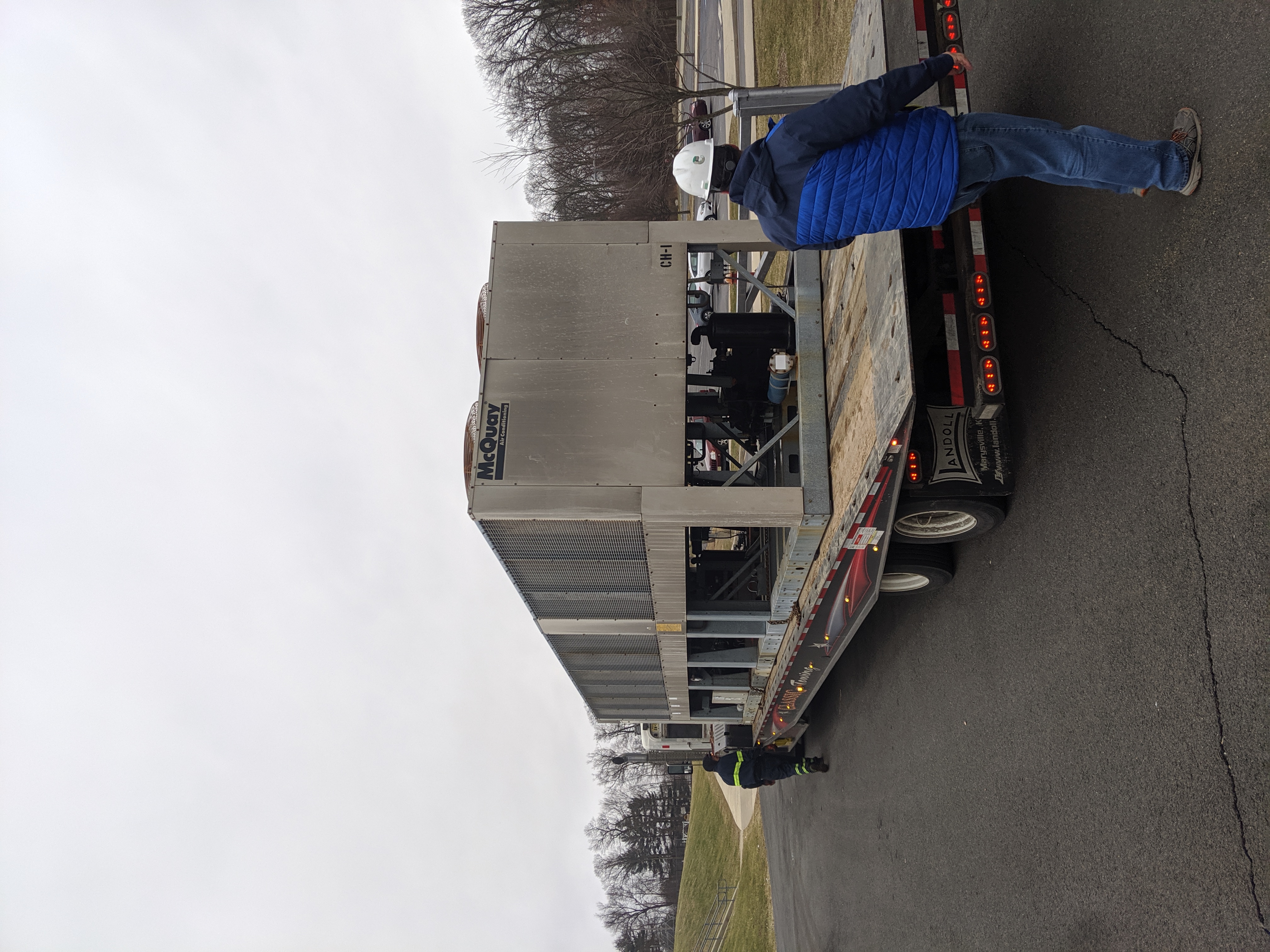 Two workers stand near a large industrial chiller unit being transported on a flatbed truck, with a treeline and overcast sky in the background.