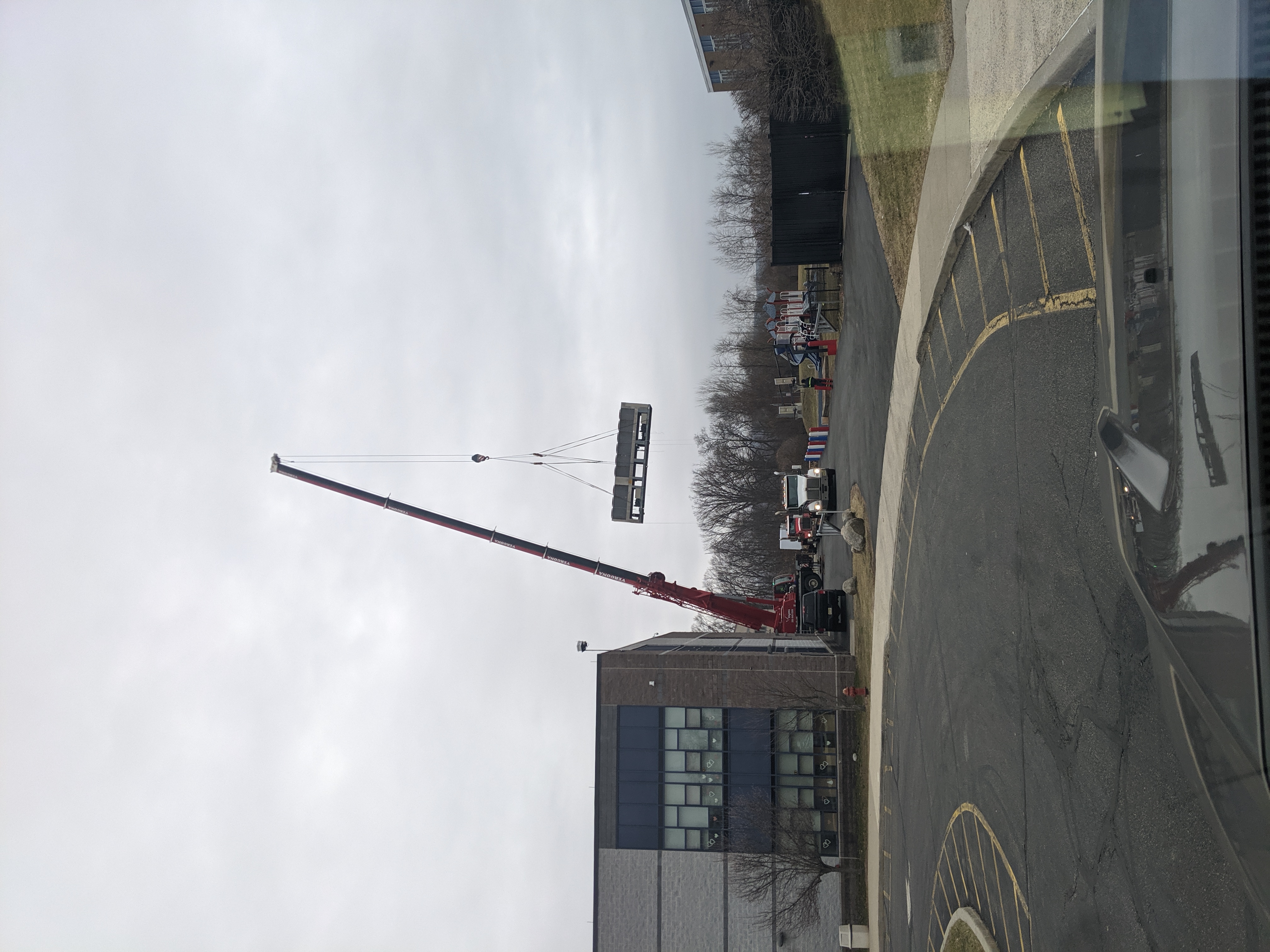A large crane lifts a rectangular object in front of a modern building, with trees and a cloudy sky in the background.