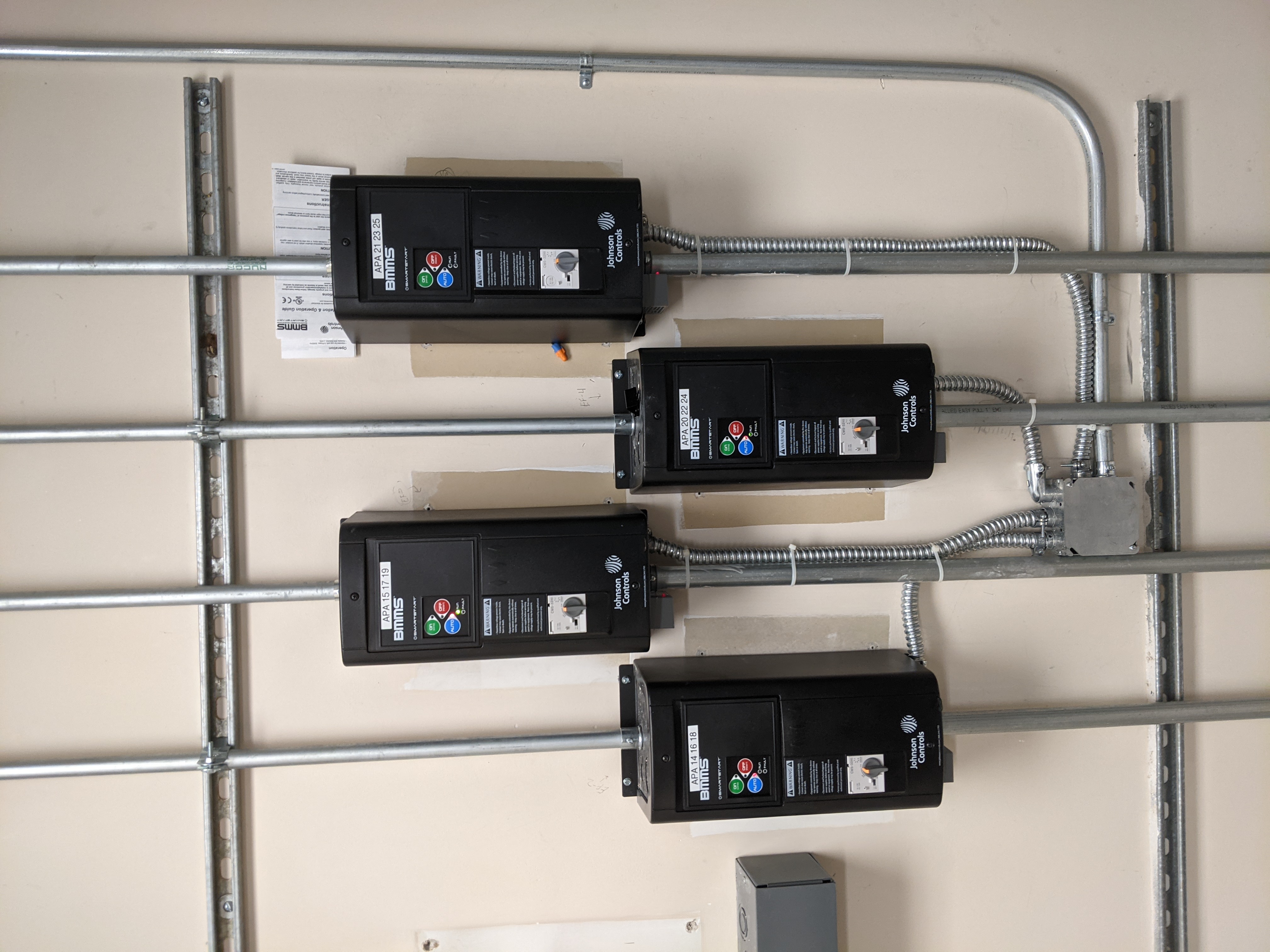 Four black electrical control boxes are mounted on a beige wall, with metal conduit pipes running vertically and horizontally in the foreground and background.