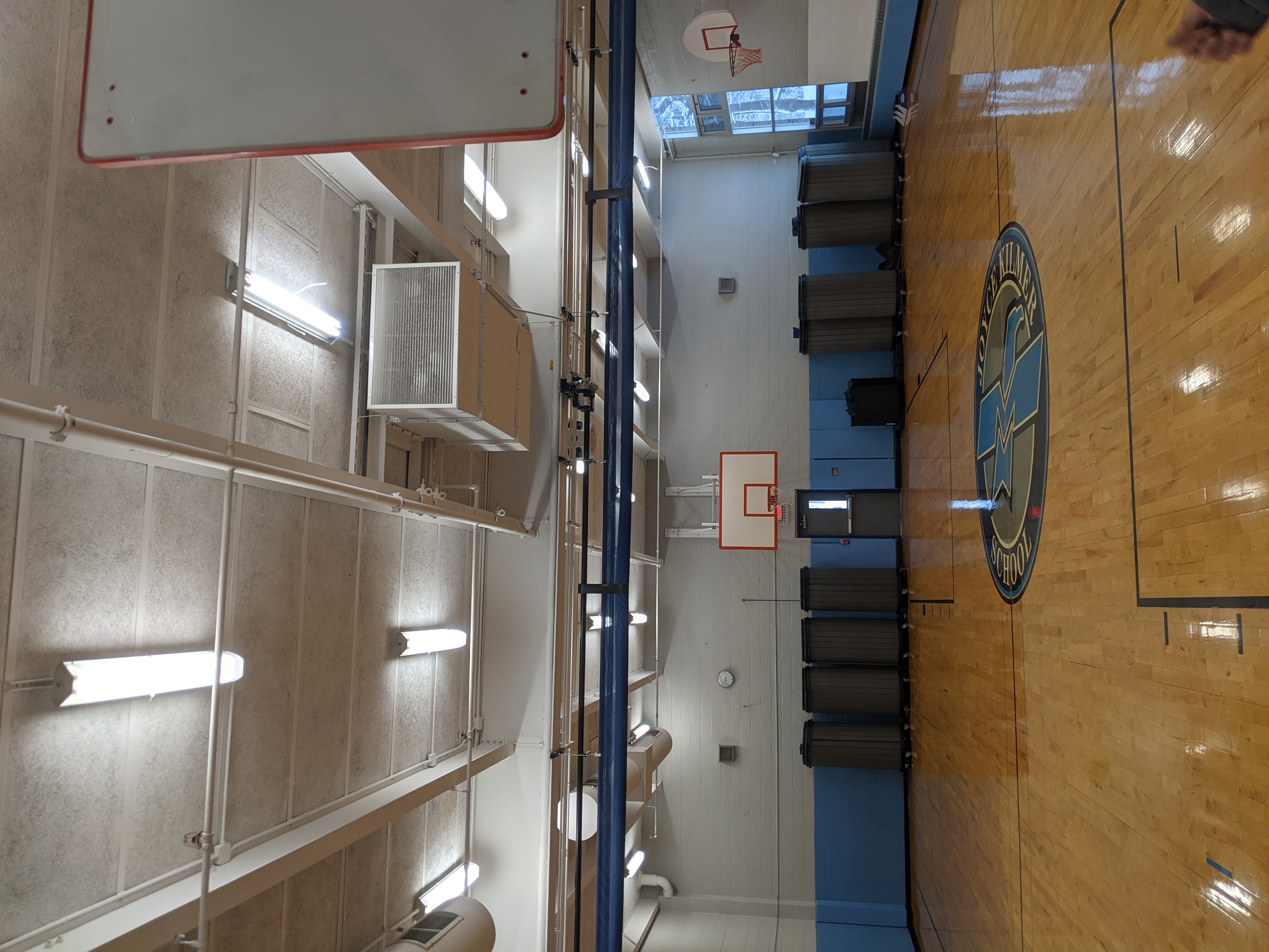 A polished wooden basketball court with a circular logo in the foreground is set against a background of gym equipment, basketball hoops, and fluorescent lighting.