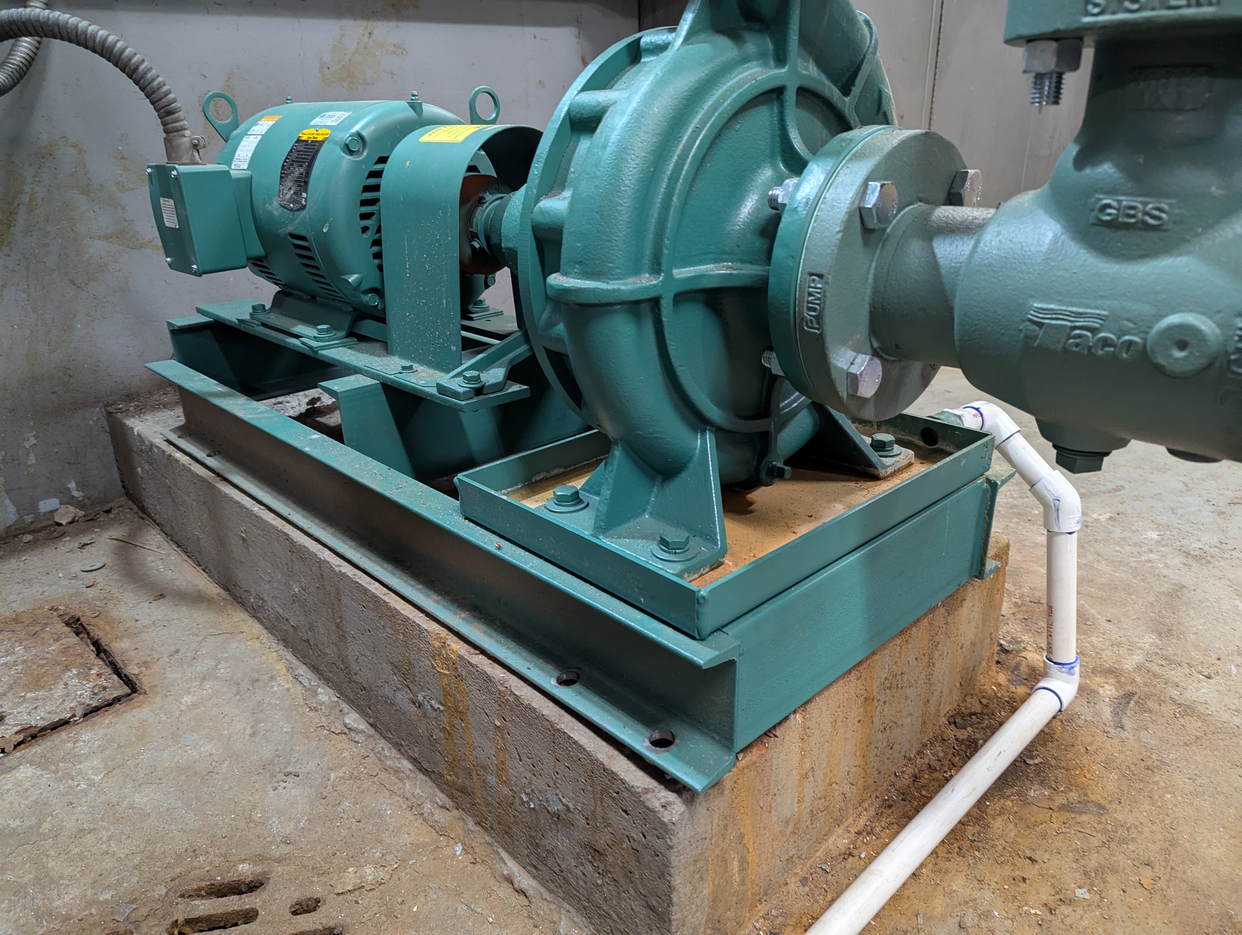 A green industrial pump and motor assembly sits on a metal base in the foreground, with a concrete floor and wall in the background.