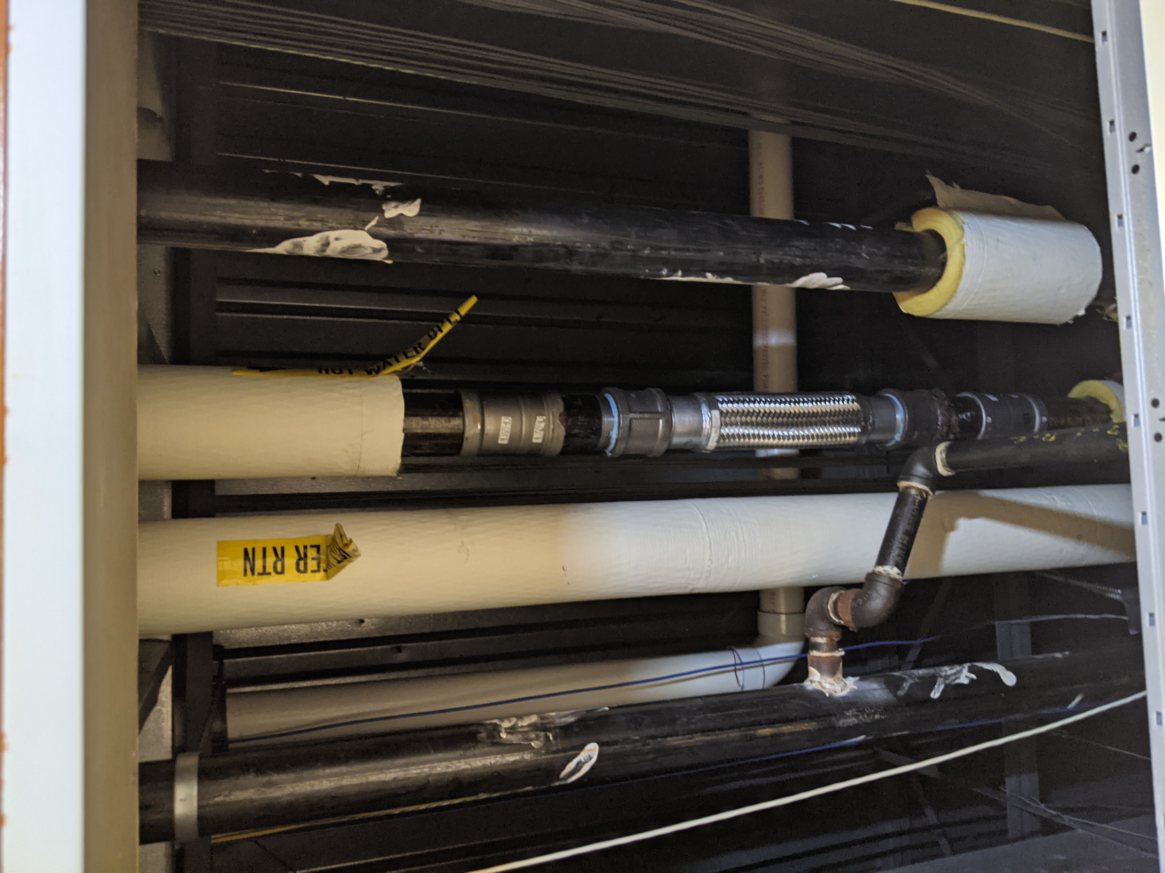 In the foreground, various pipes, including a flexible metal pipe and a black pipe with insulation, are visible, while the background shows a corrugated metal wall.