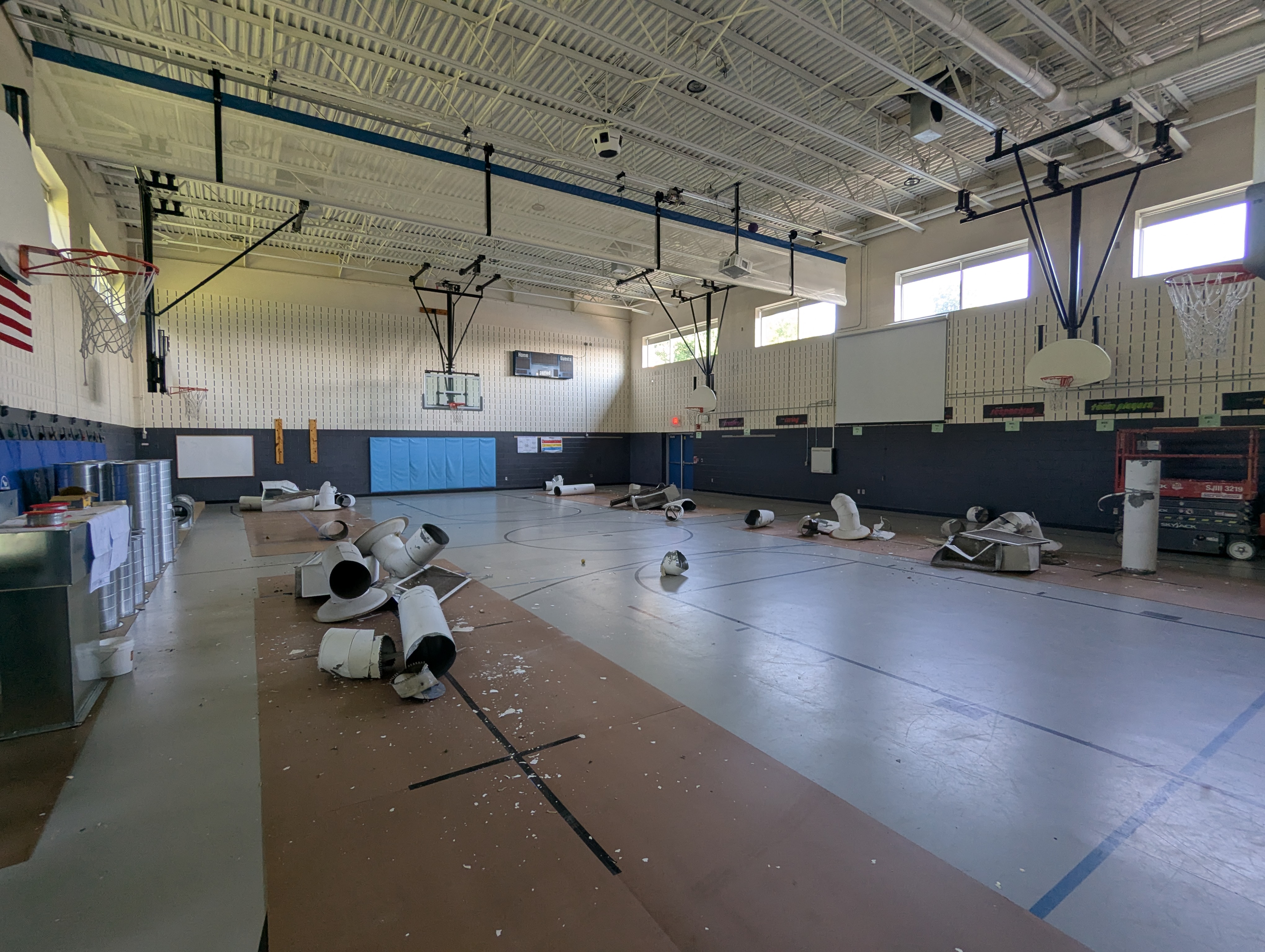In the foreground, large white cylindrical objects and debris are scattered across a brown tarp on the gym floor, while the background reveals a large, empty gymnasium with basketball hoops and windows.