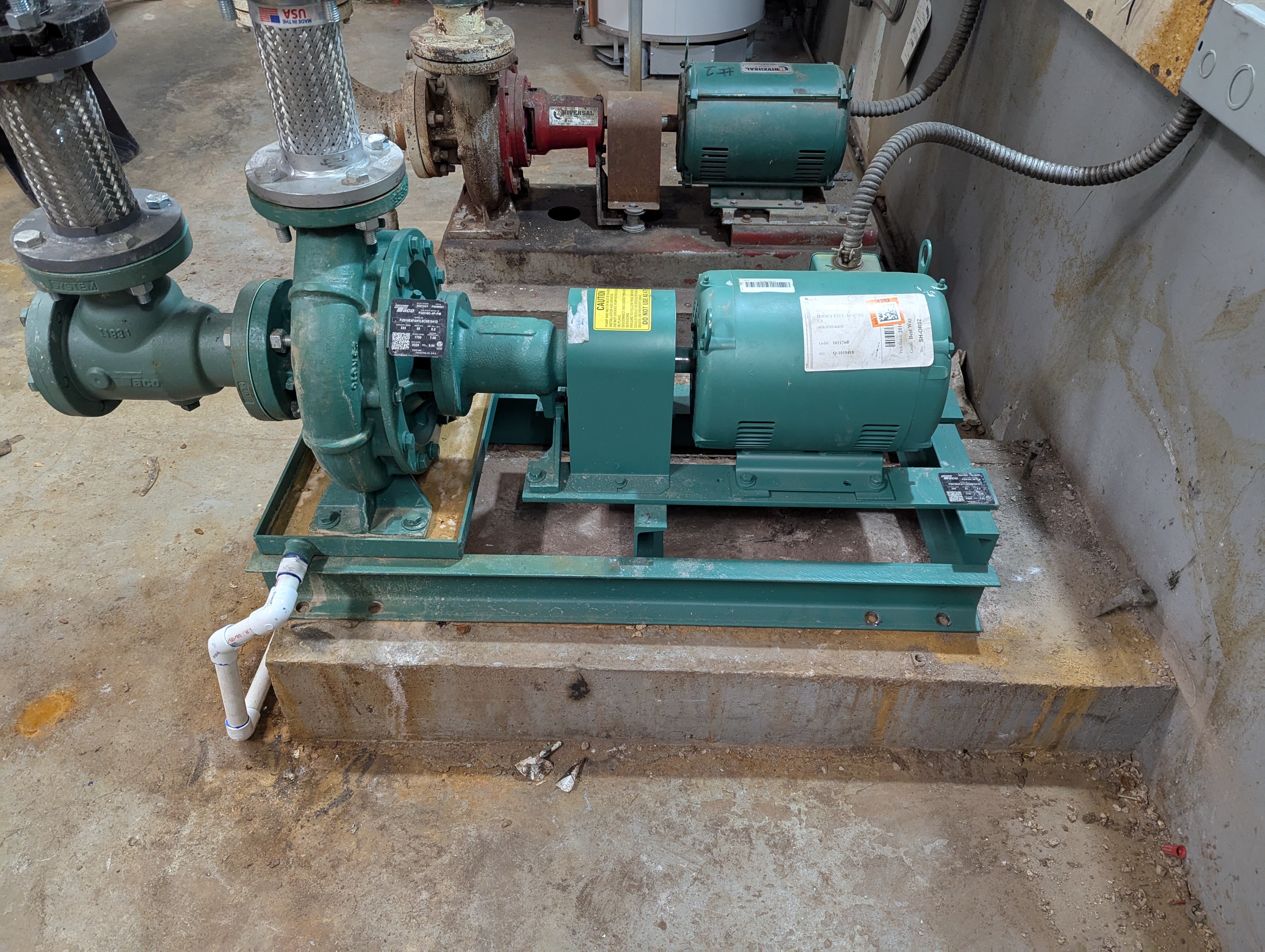 A green industrial pump with a motor sits on a metal base in the foreground, with other machinery and pipes visible in the background.