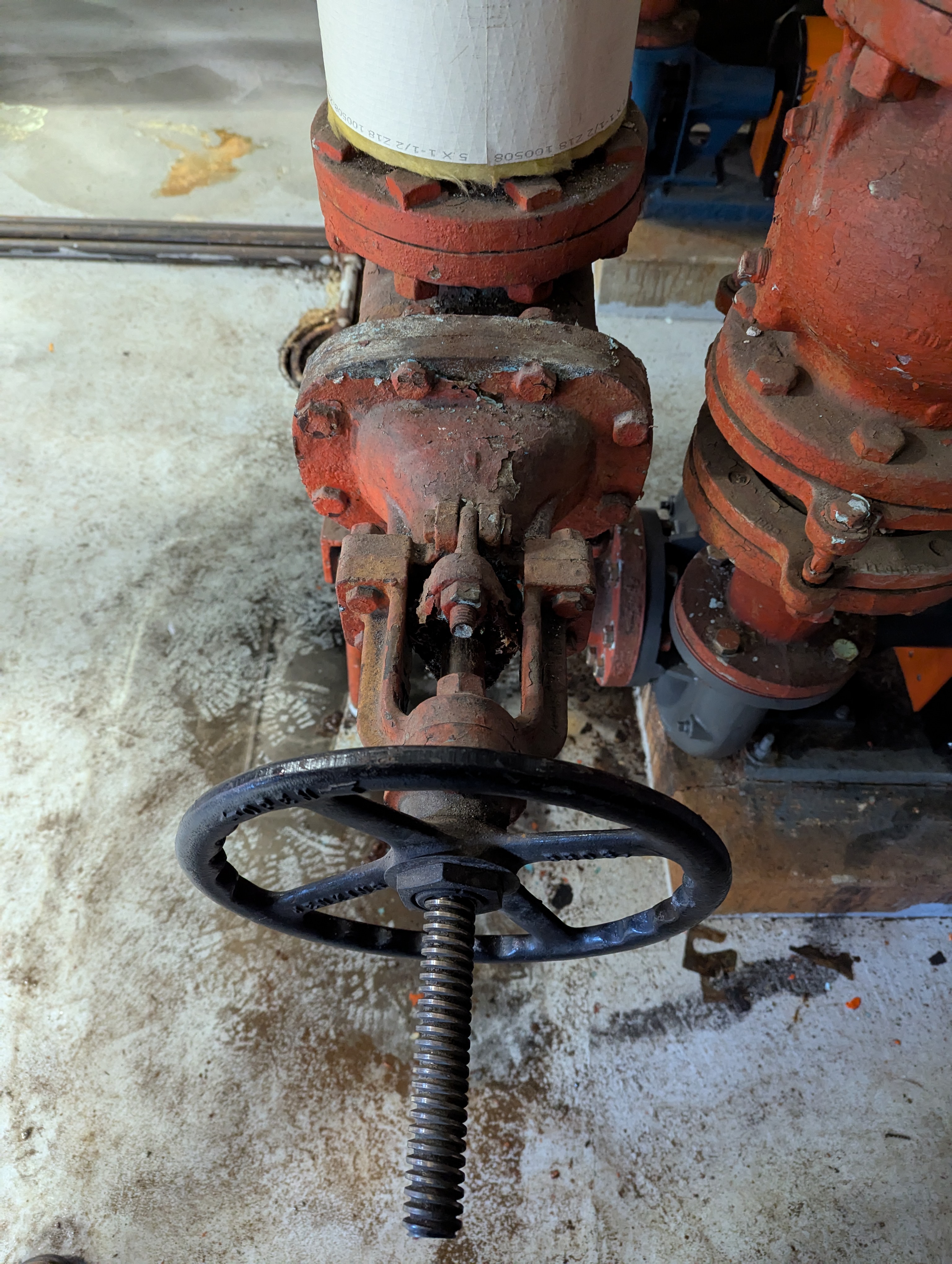 A large, rusty valve with a black handwheel is in the foreground, with industrial pipes and machinery visible in the background.