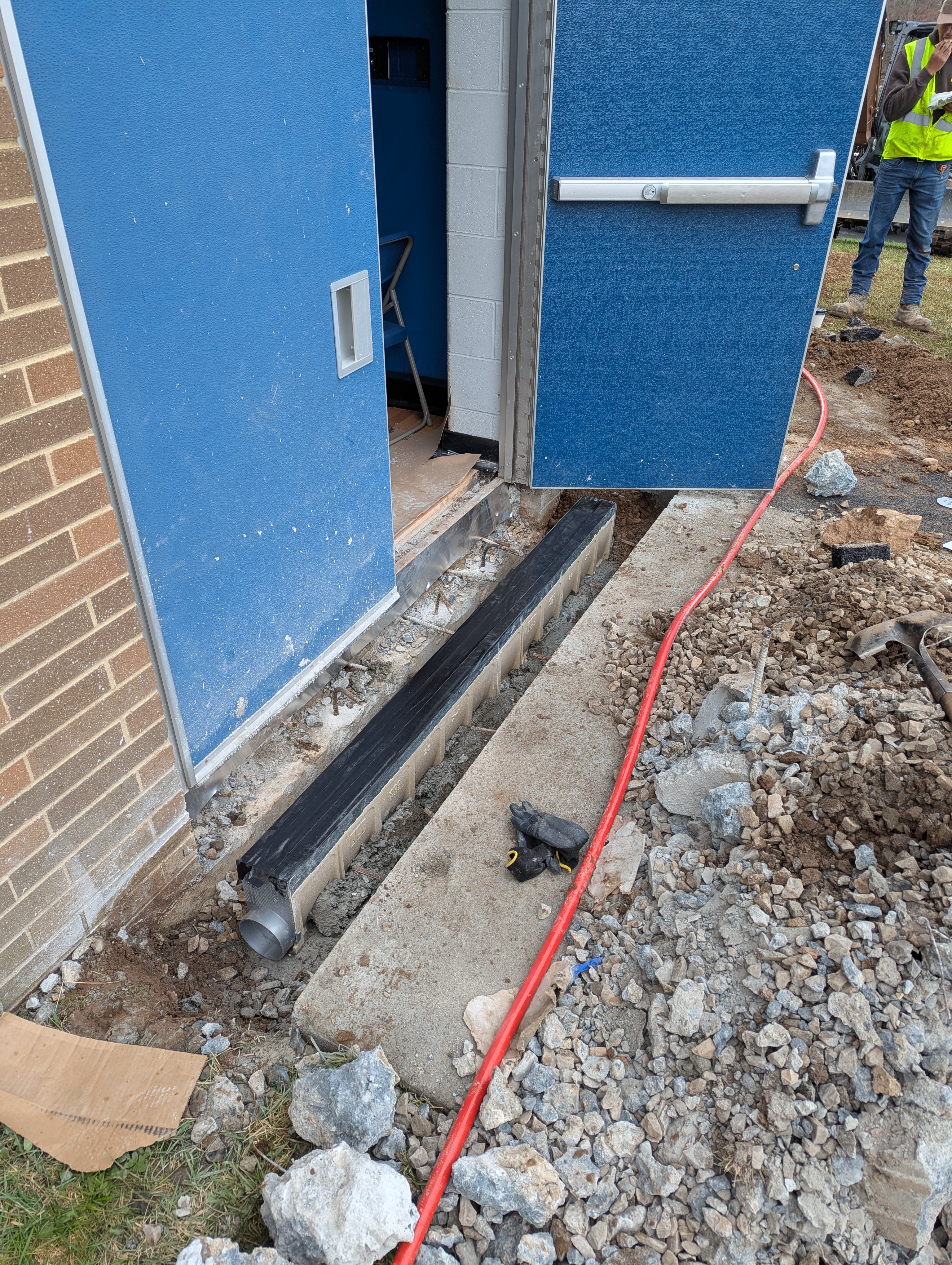 A drainage grate is installed in front of a blue door, with construction debris and a red cable scattered around the foreground and a person in a safety vest visible in the background.