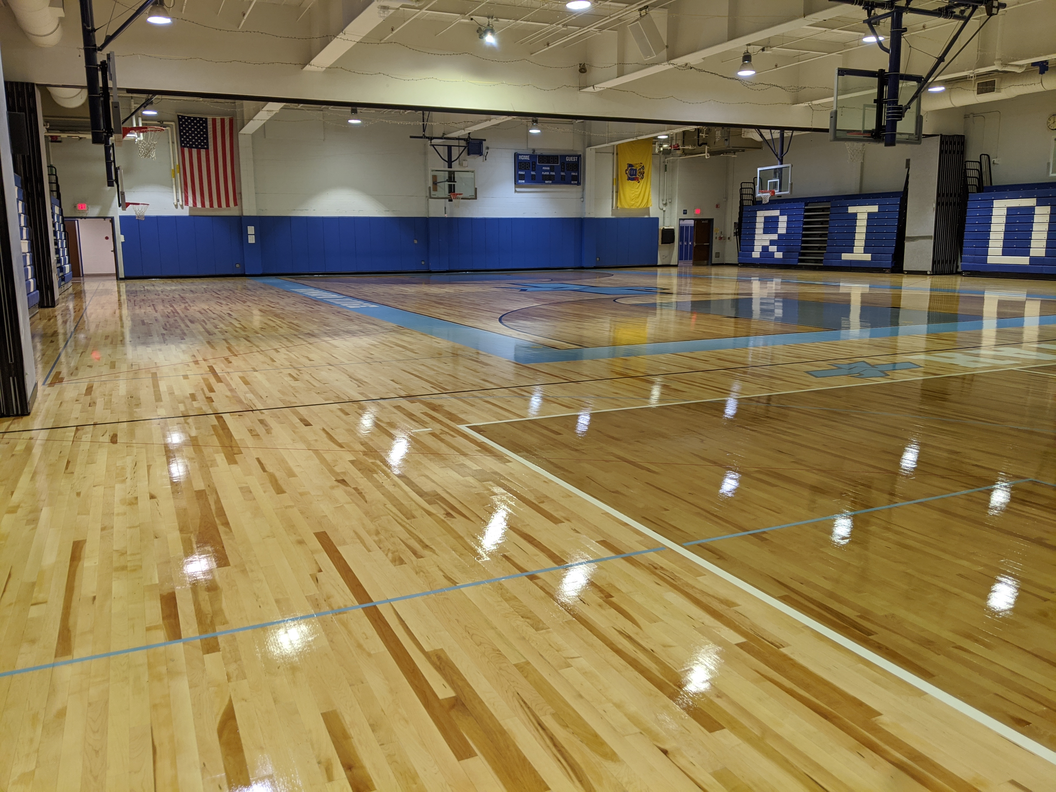 A polished wooden basketball court with blue accents fills the foreground, while the background reveals bleachers, an American flag, and a scoreboard in a gymnasium.