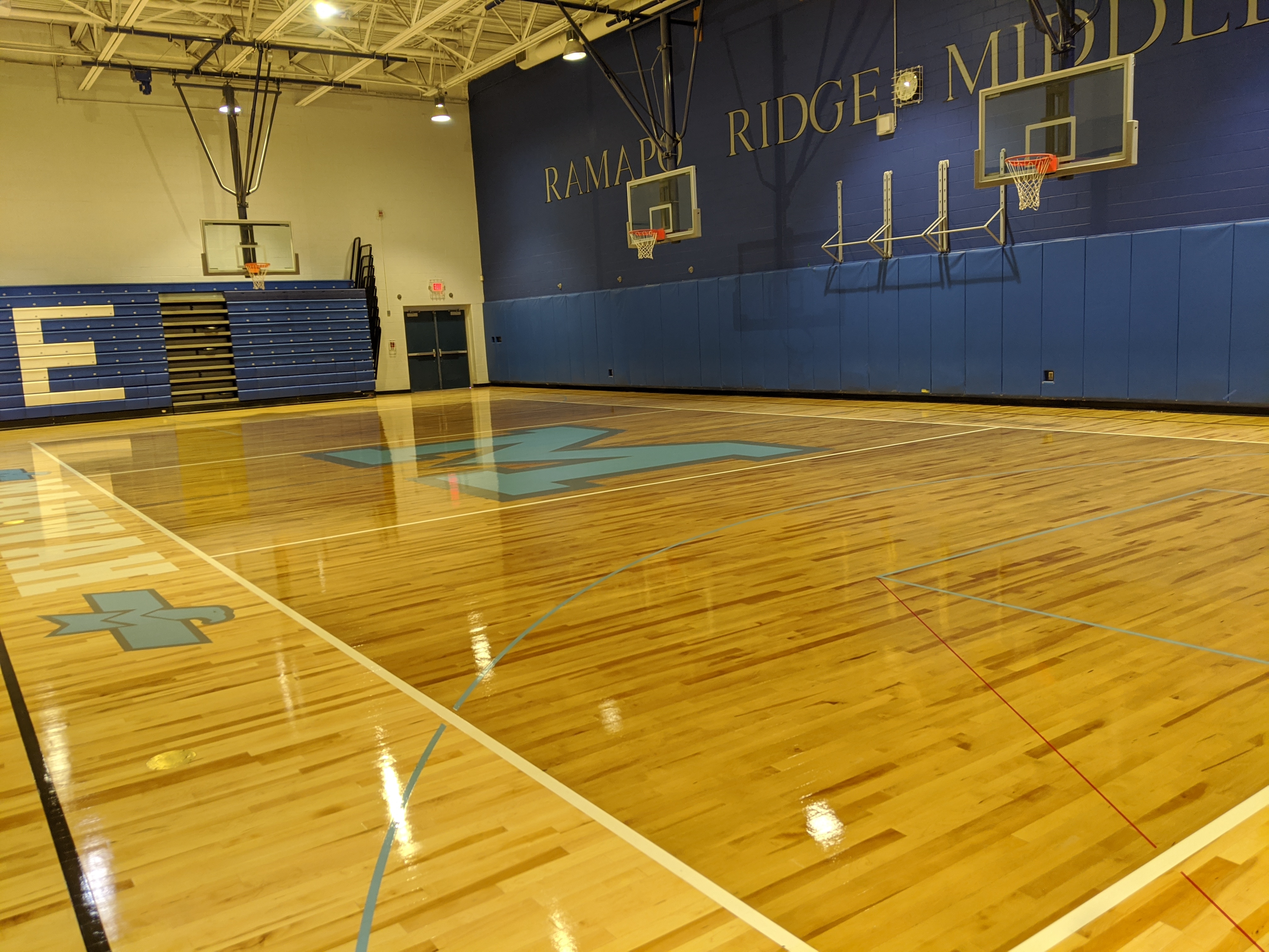 A polished wooden basketball court with blue markings fills the foreground, while bleachers and a blue wall with "RAMAPO RIDGE MIDDLE" lettering and basketball hoops form the background.