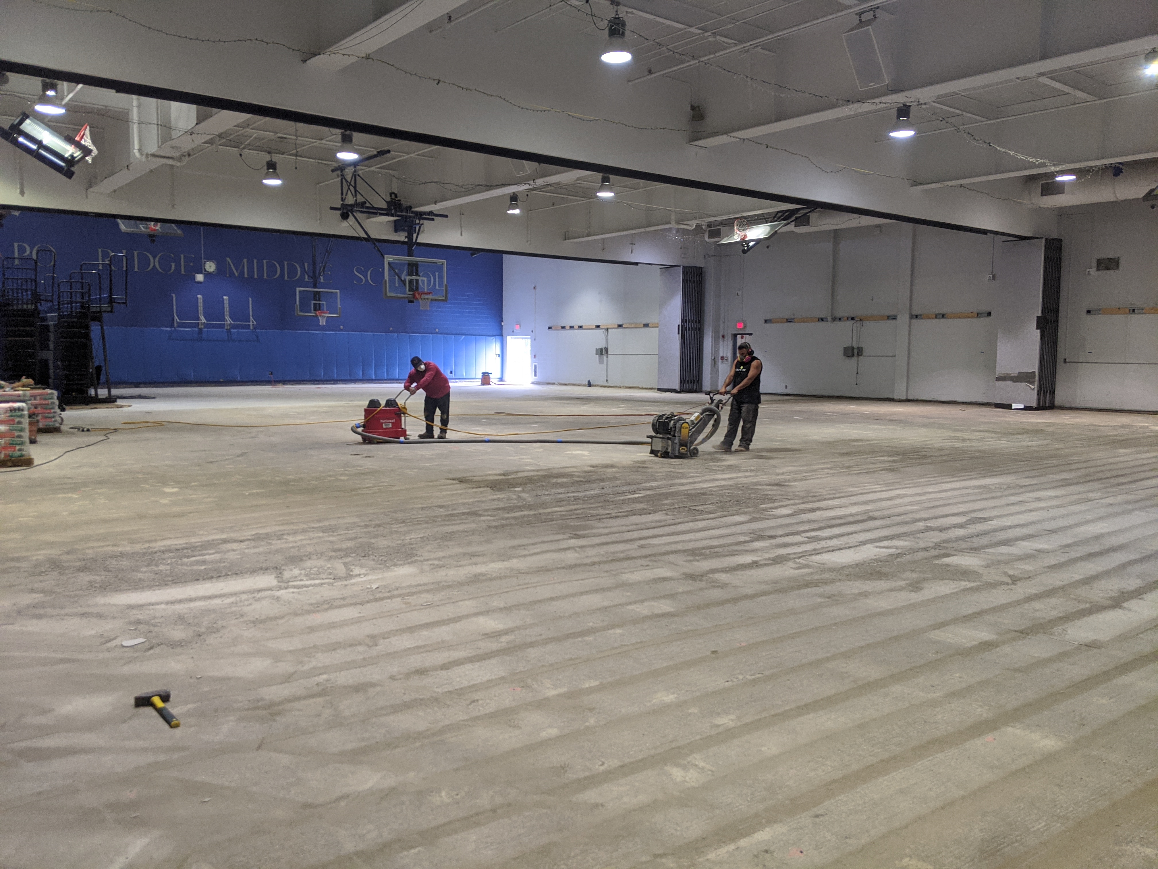 Two workers are grinding a large, unfinished concrete floor in a gymnasium with a blue wall and basketball hoops in the background.