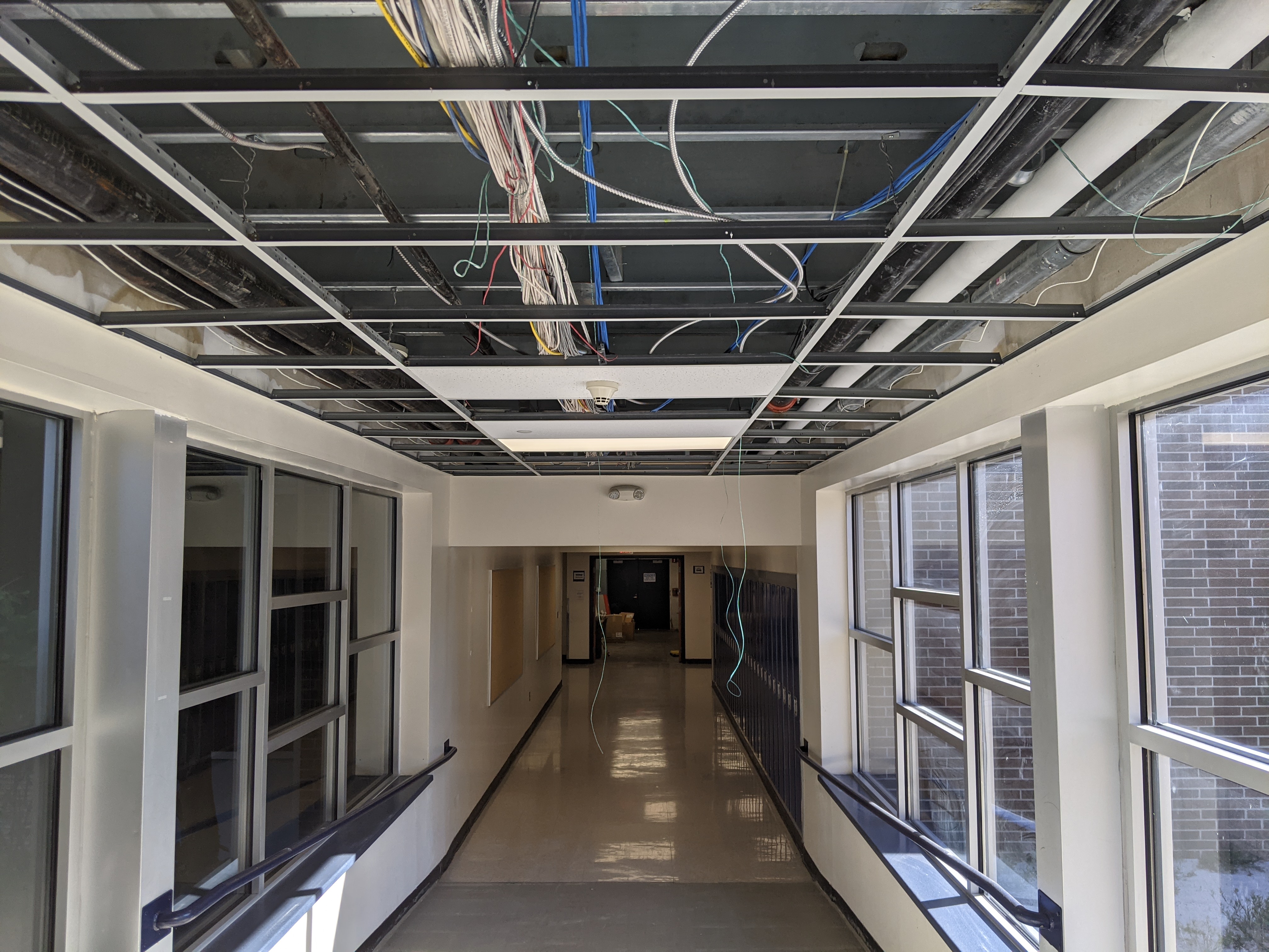 Exposed wiring and pipes are visible in the open ceiling of a hallway with large windows on either side, leading to a dimly lit corridor in the background.