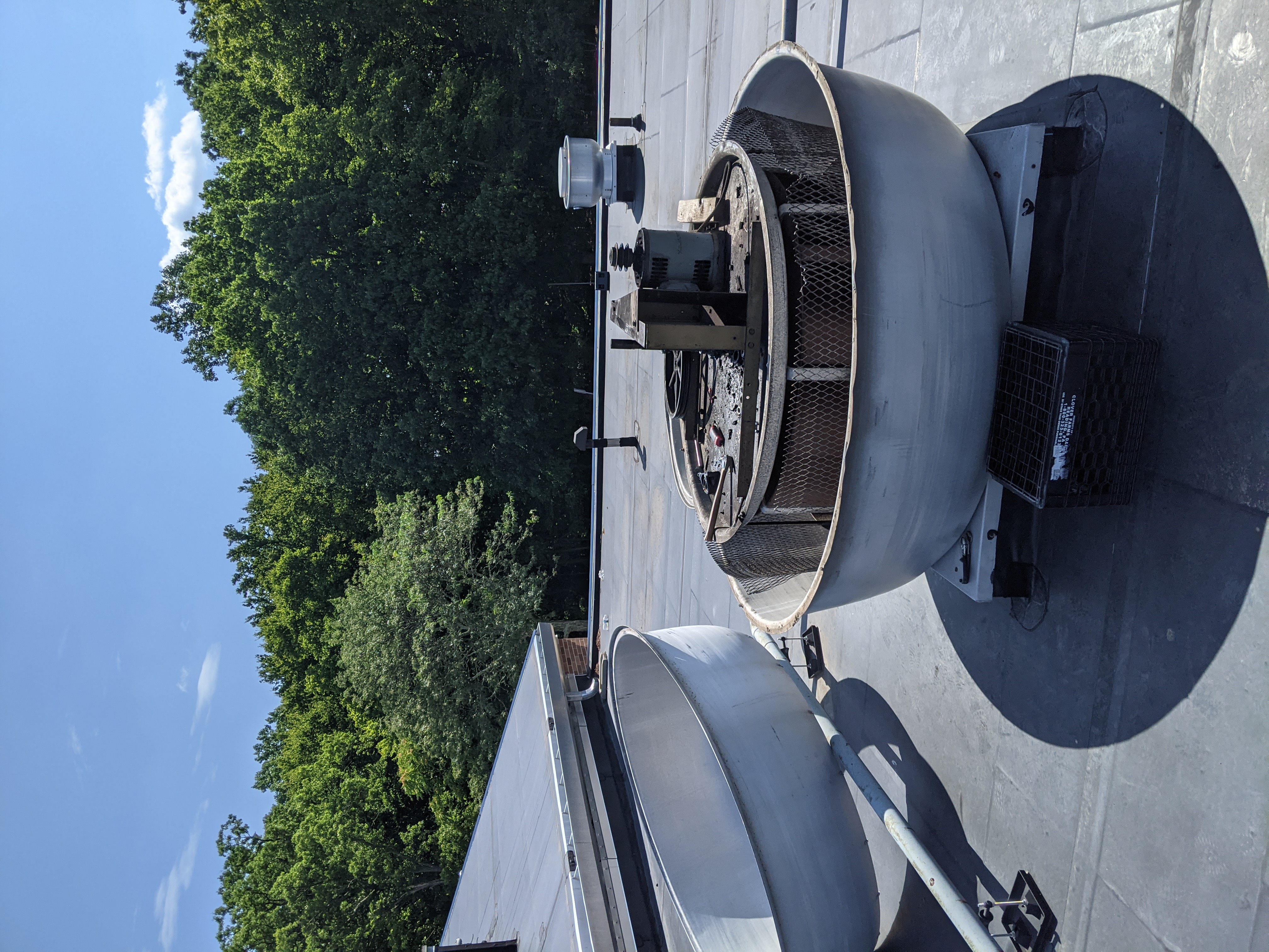 Two large, grey, industrial-looking basins sit on a dark, flat rooftop, with a dense green forest and a bright blue sky in the background.