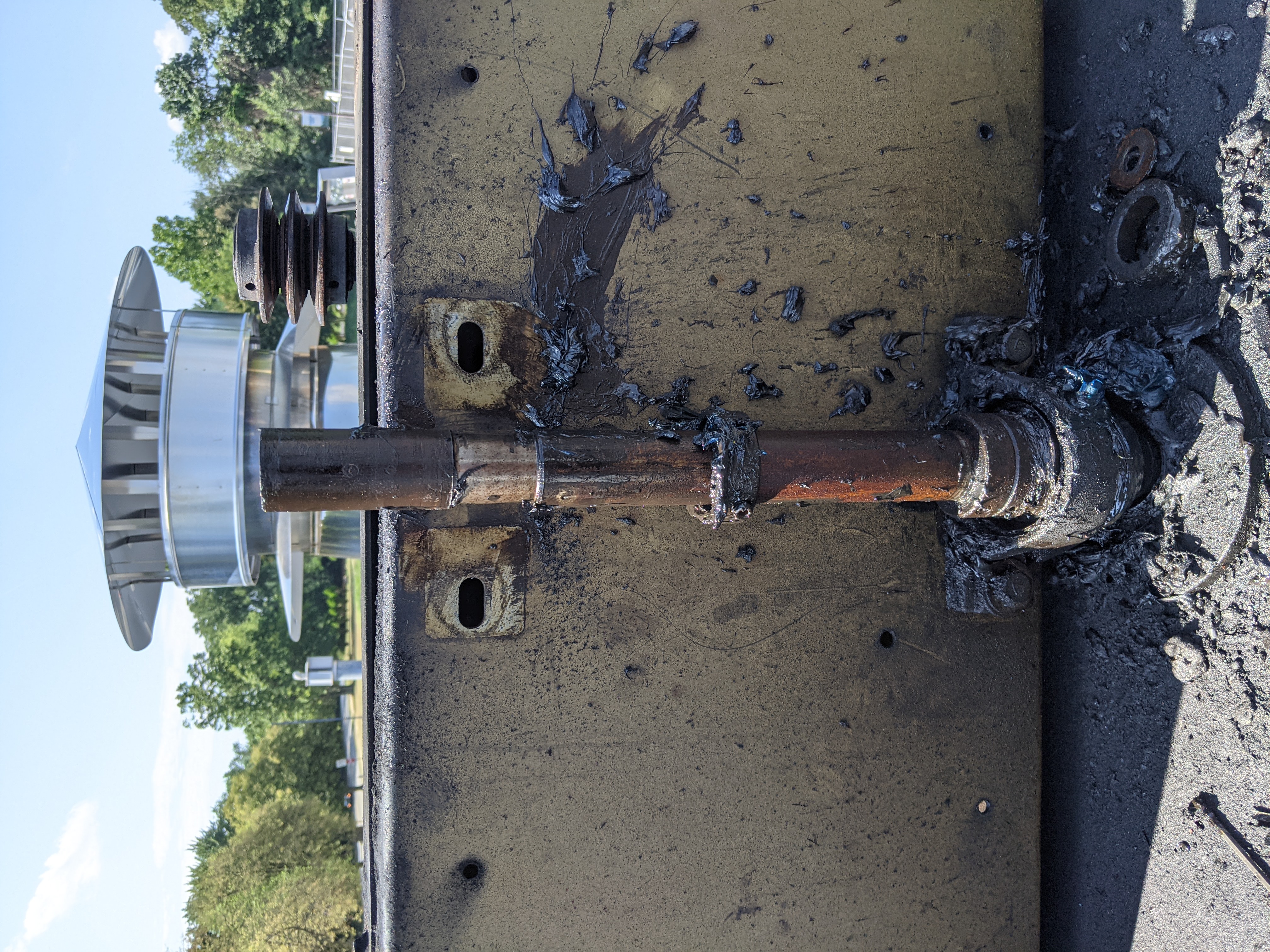 A rusty pipe protrudes from a weathered surface in the foreground, with a shiny vent cap and trees visible in the background.