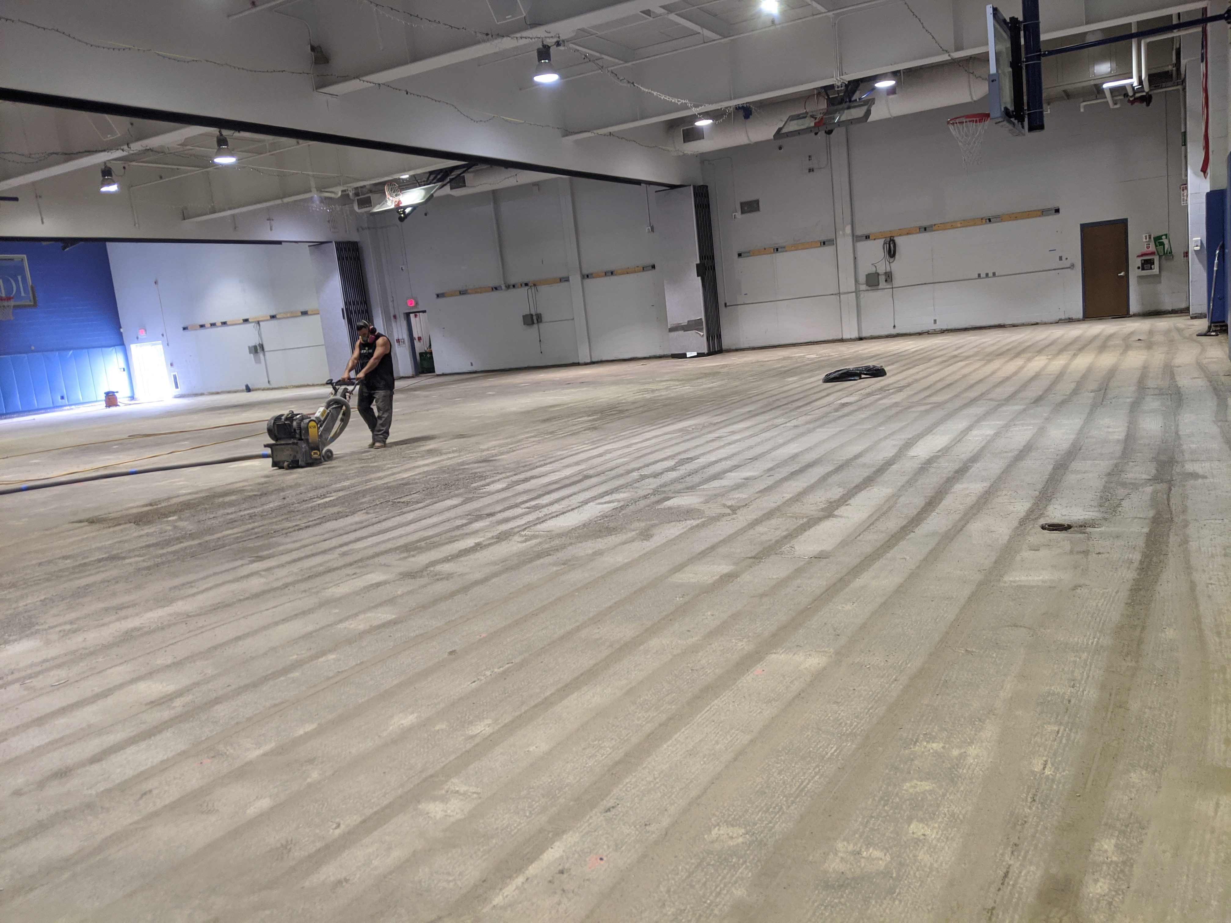 A person operates a floor grinder on a vast, newly prepped concrete floor in a large gymnasium with white walls and garage doors in the background.