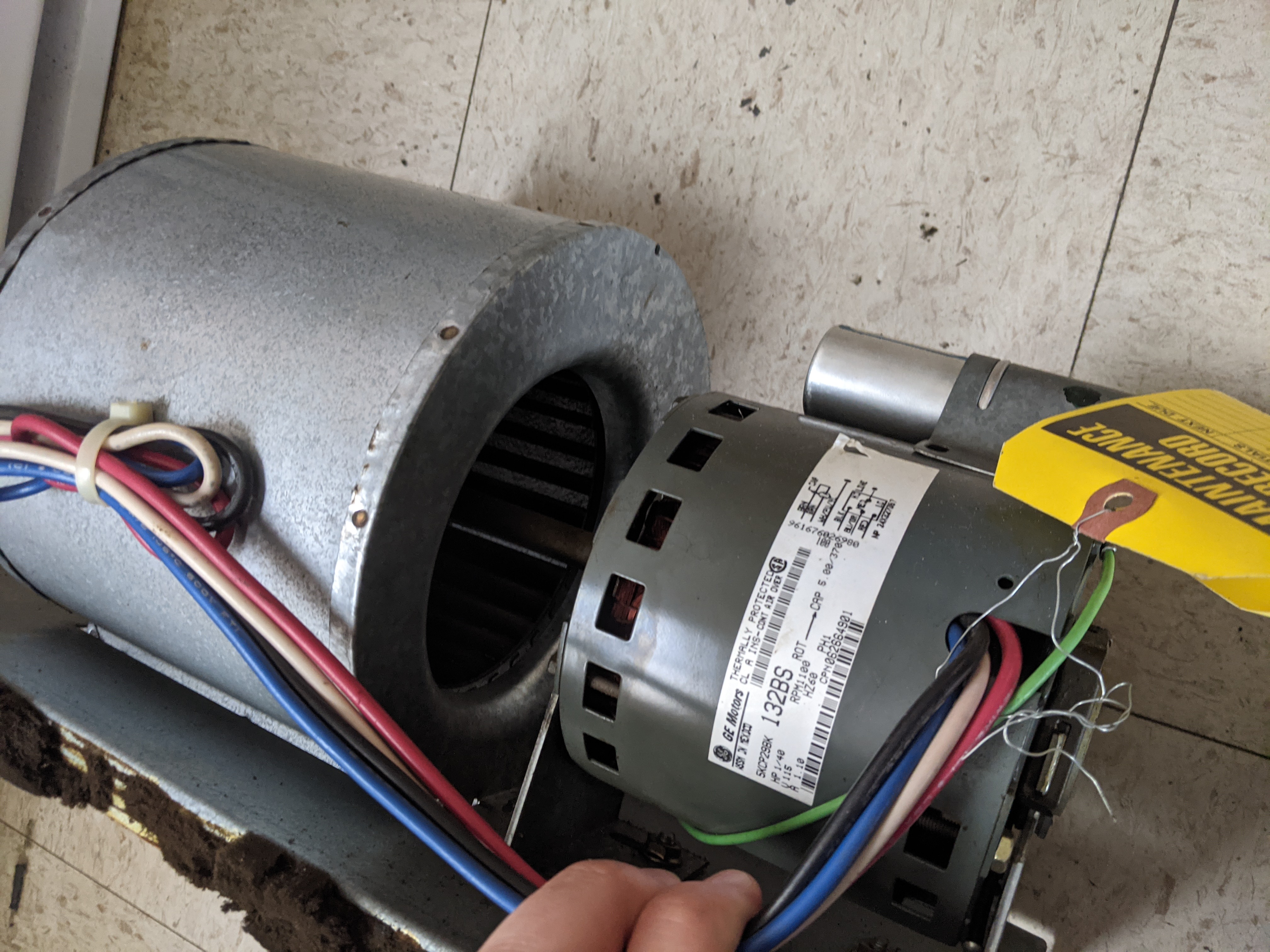 A hand holds wires connected to a motor and blower assembly in the foreground, with a white appliance visible in the background.