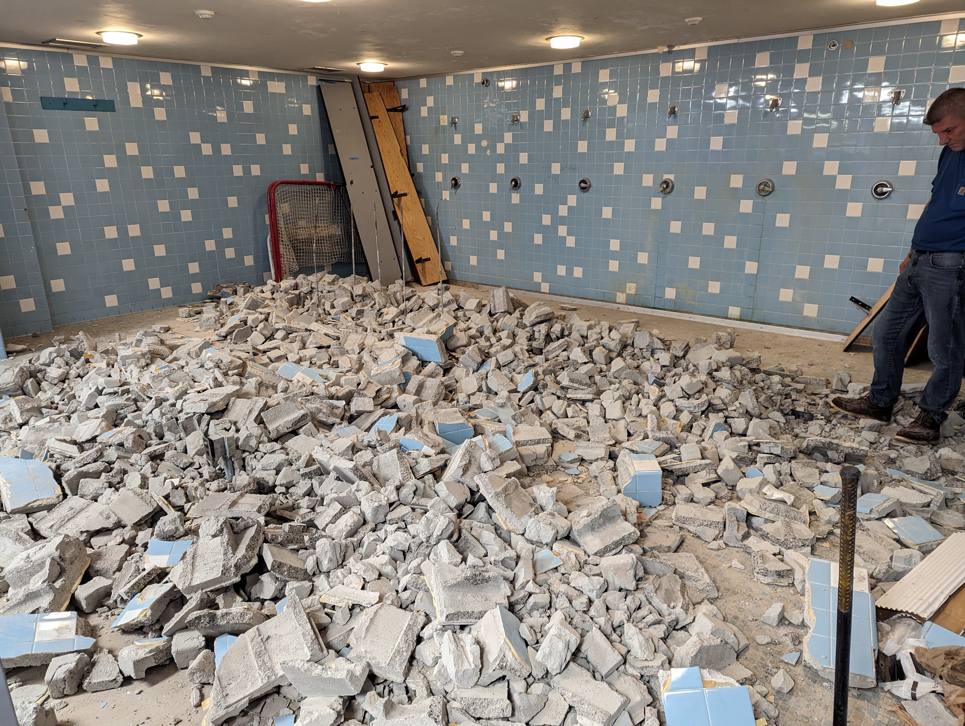 A man stands near a pile of rubble in a room with blue tiled walls and showers.