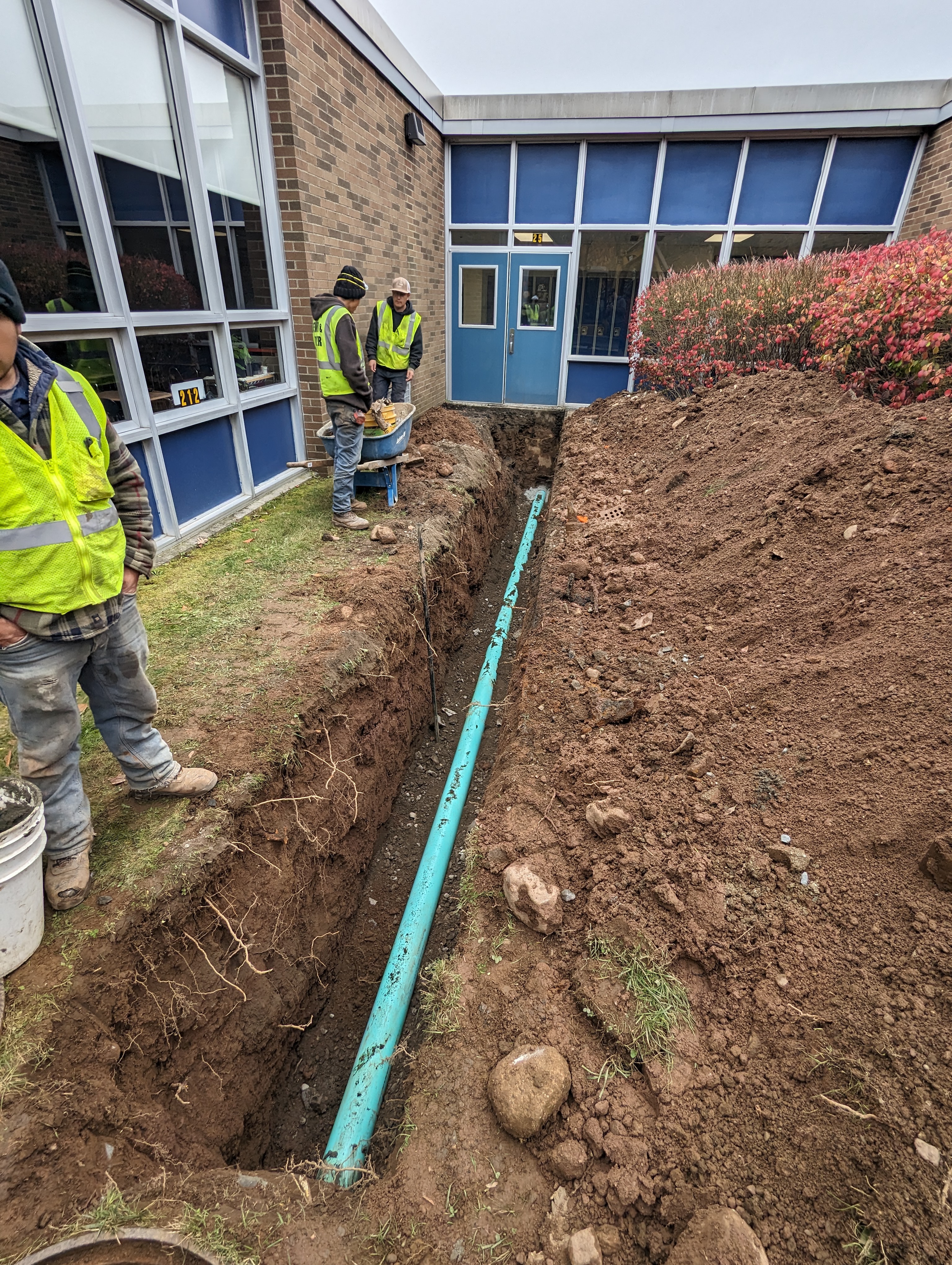 Workers in high-visibility vests stand around a deep trench with a turquoise pipe, while a building with blue-trimmed windows forms the background.