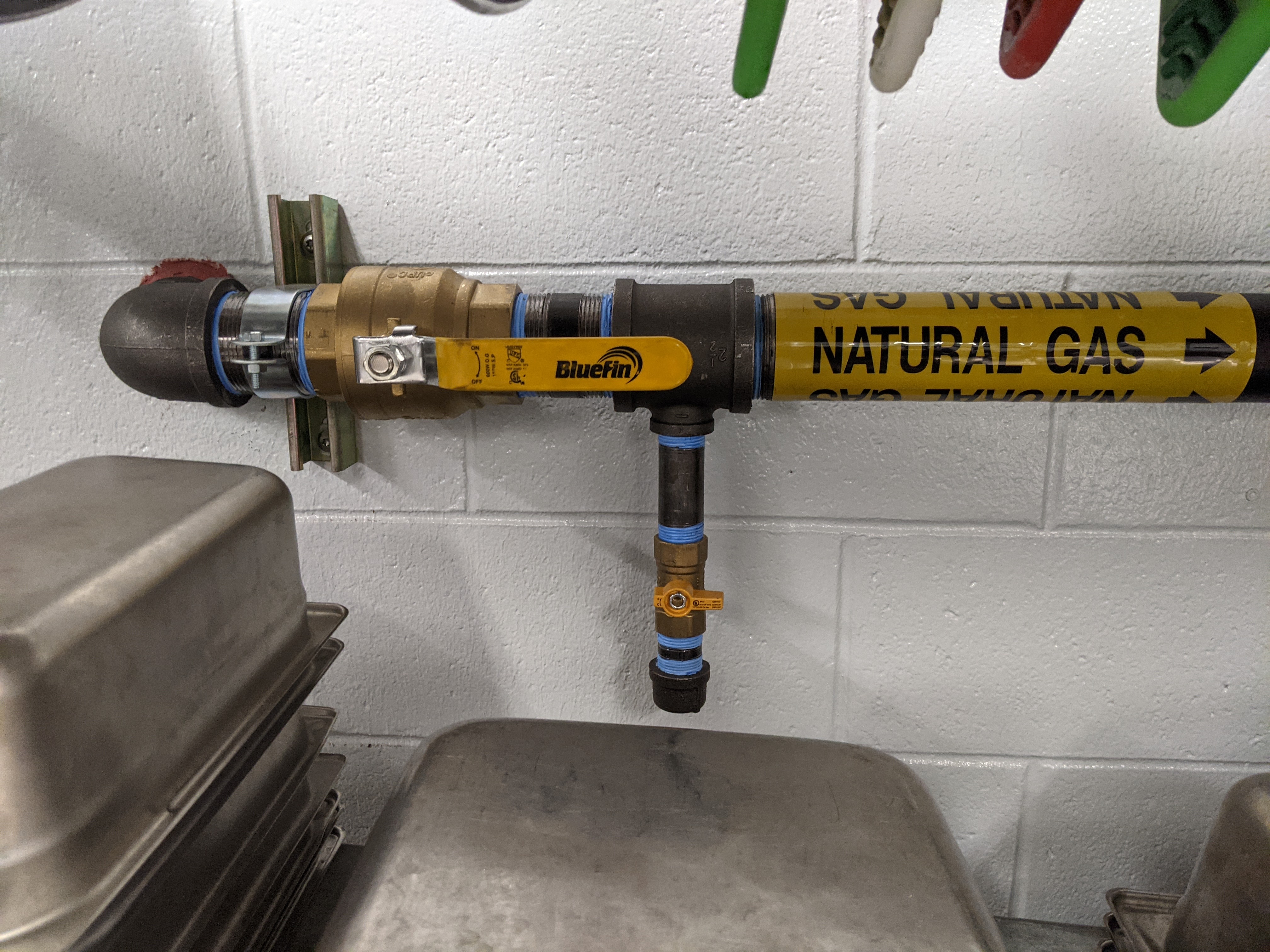 In the foreground, stacked metal trays sit below a natural gas pipe with a yellow valve, while the background features a white brick wall.