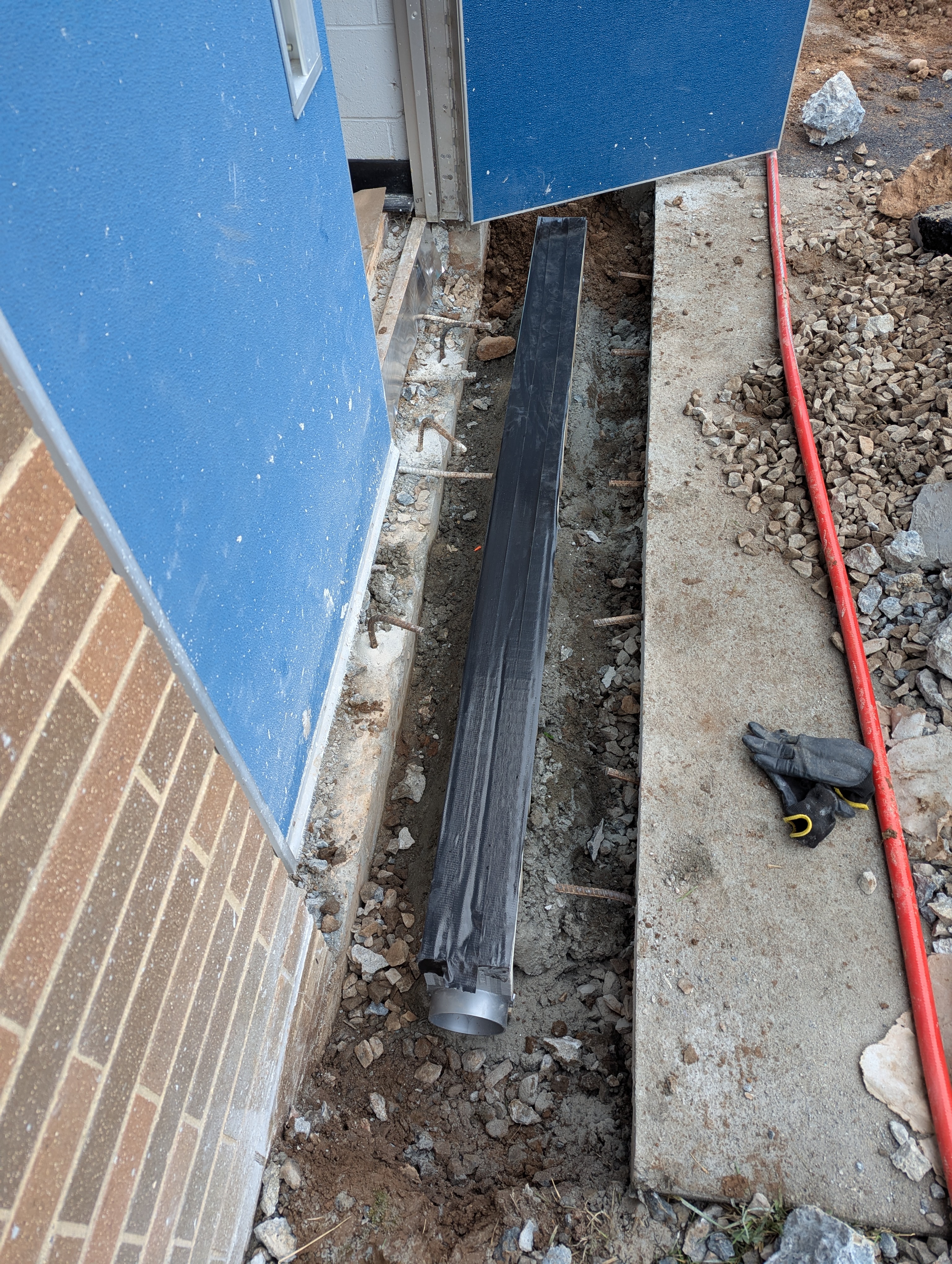 A black drainage pipe lies in a trench in the foreground, with a blue wall and brickwork to the left and a concrete path and gravel to the right in the background.