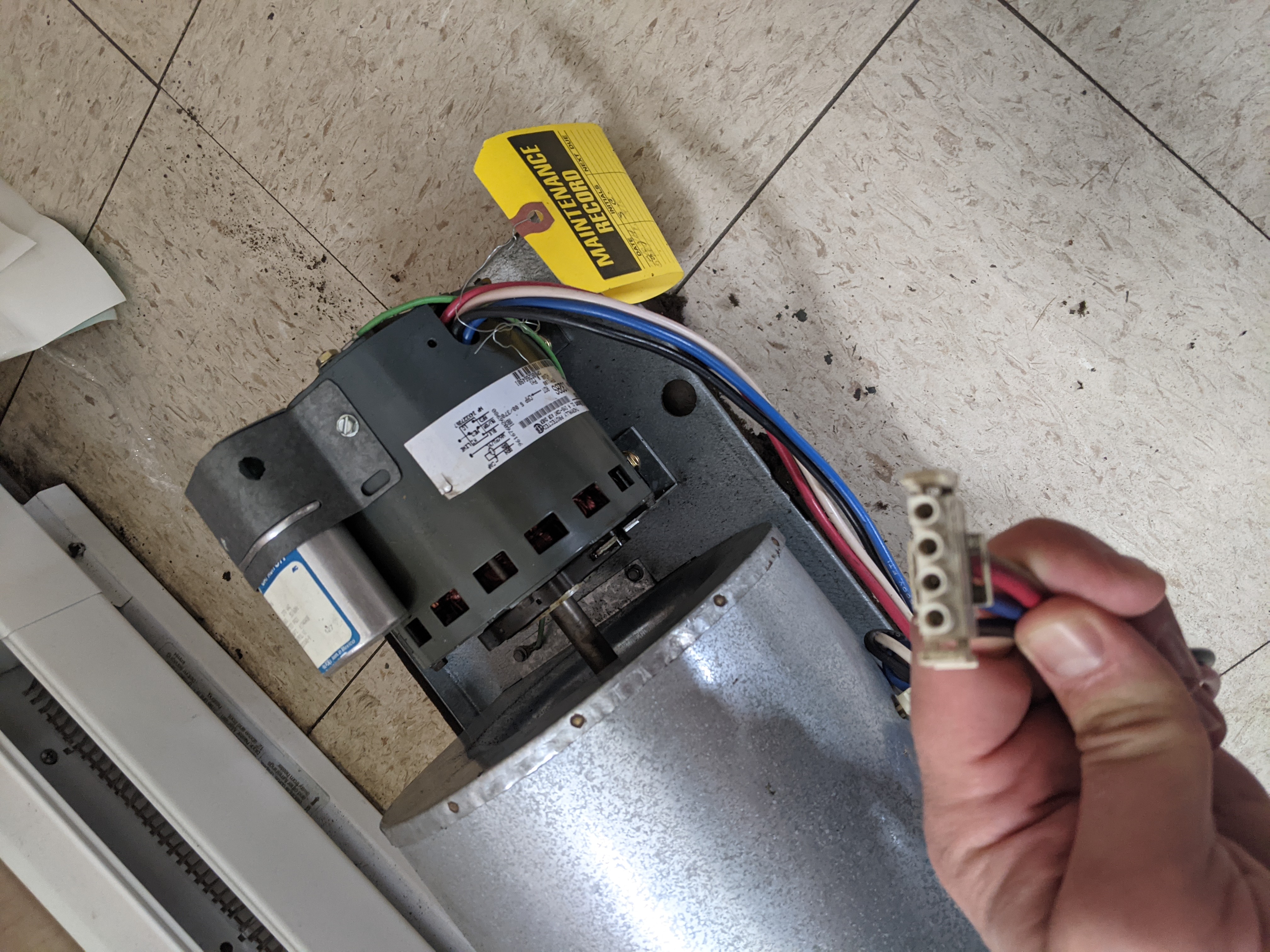 A hand holds a disconnected electrical plug in the foreground, with a furnace blower motor and a yellow maintenance tag visible in the background.