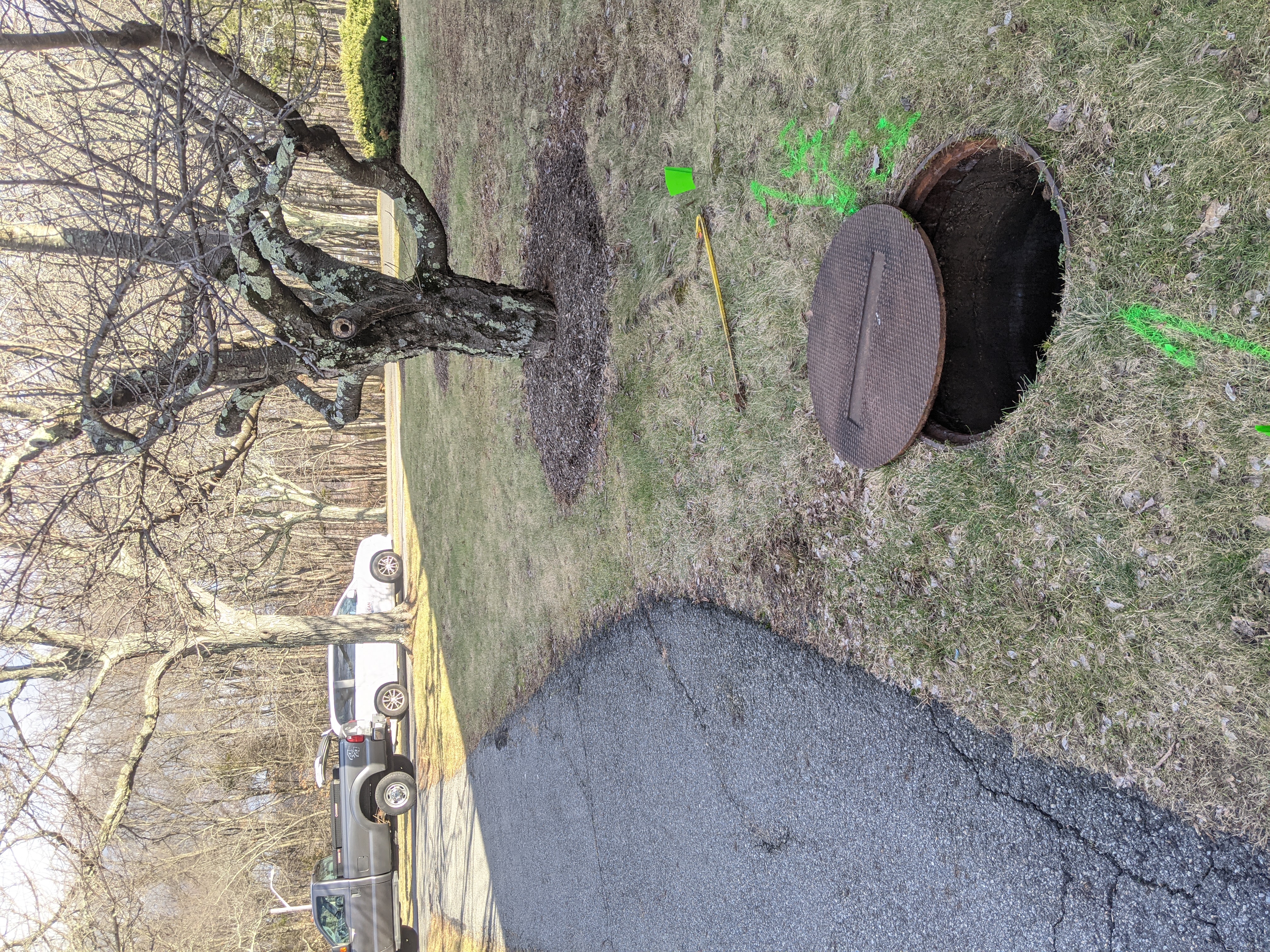 A rusty manhole cover lies open next to a tree on a grassy lawn, with a paved driveway and parked vehicles in the background.