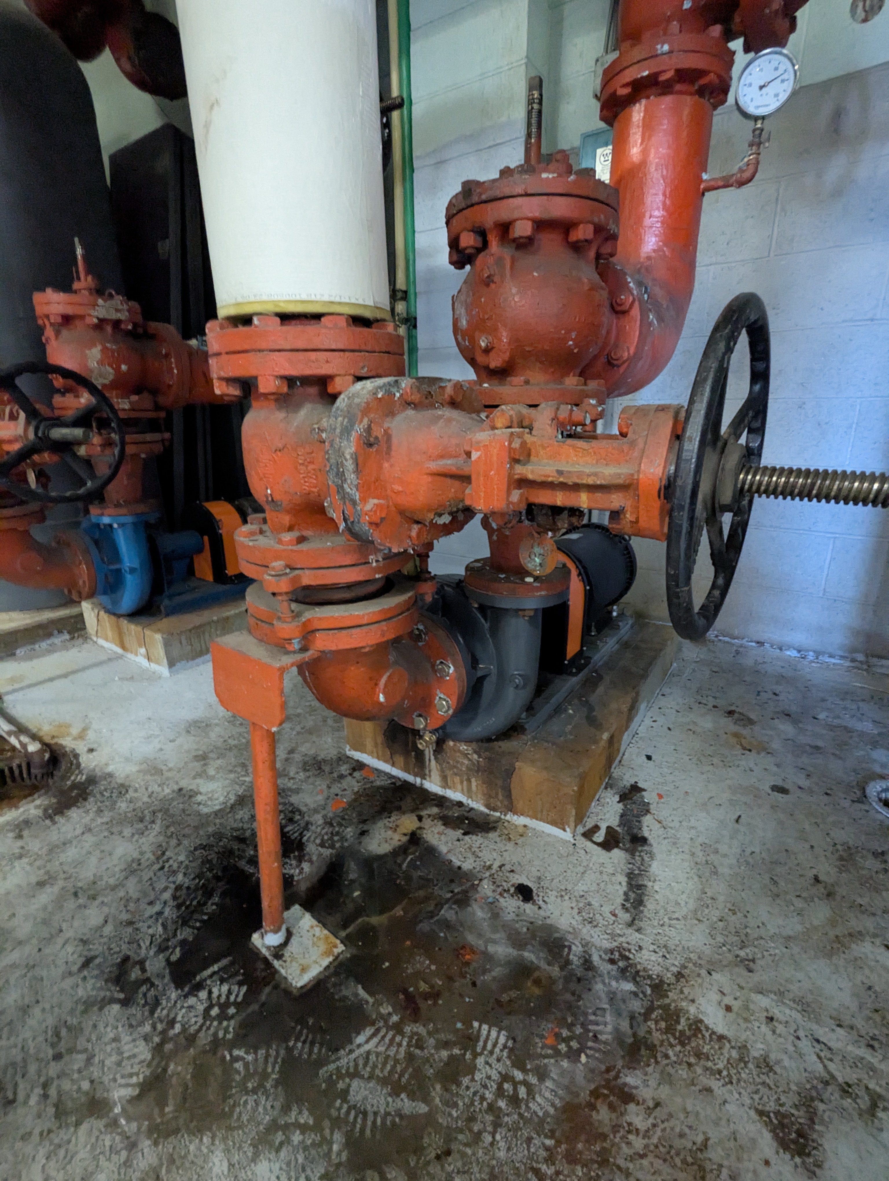In the foreground, a rusty orange pump with a large wheel sits on wooden blocks, while the background features pipes and a white cylindrical tank.