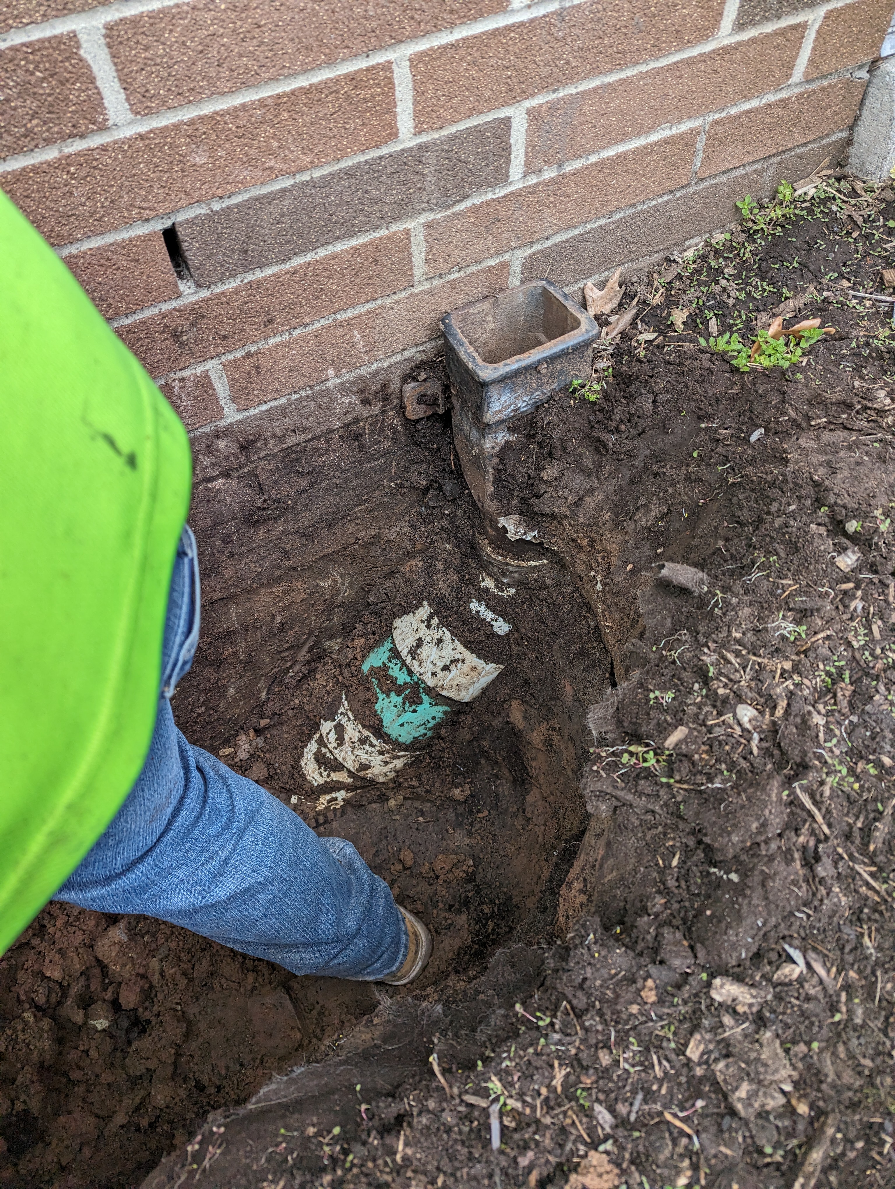 A person in a neon green shirt and blue jeans works in a trench exposing a broken pipe next to a brick wall.