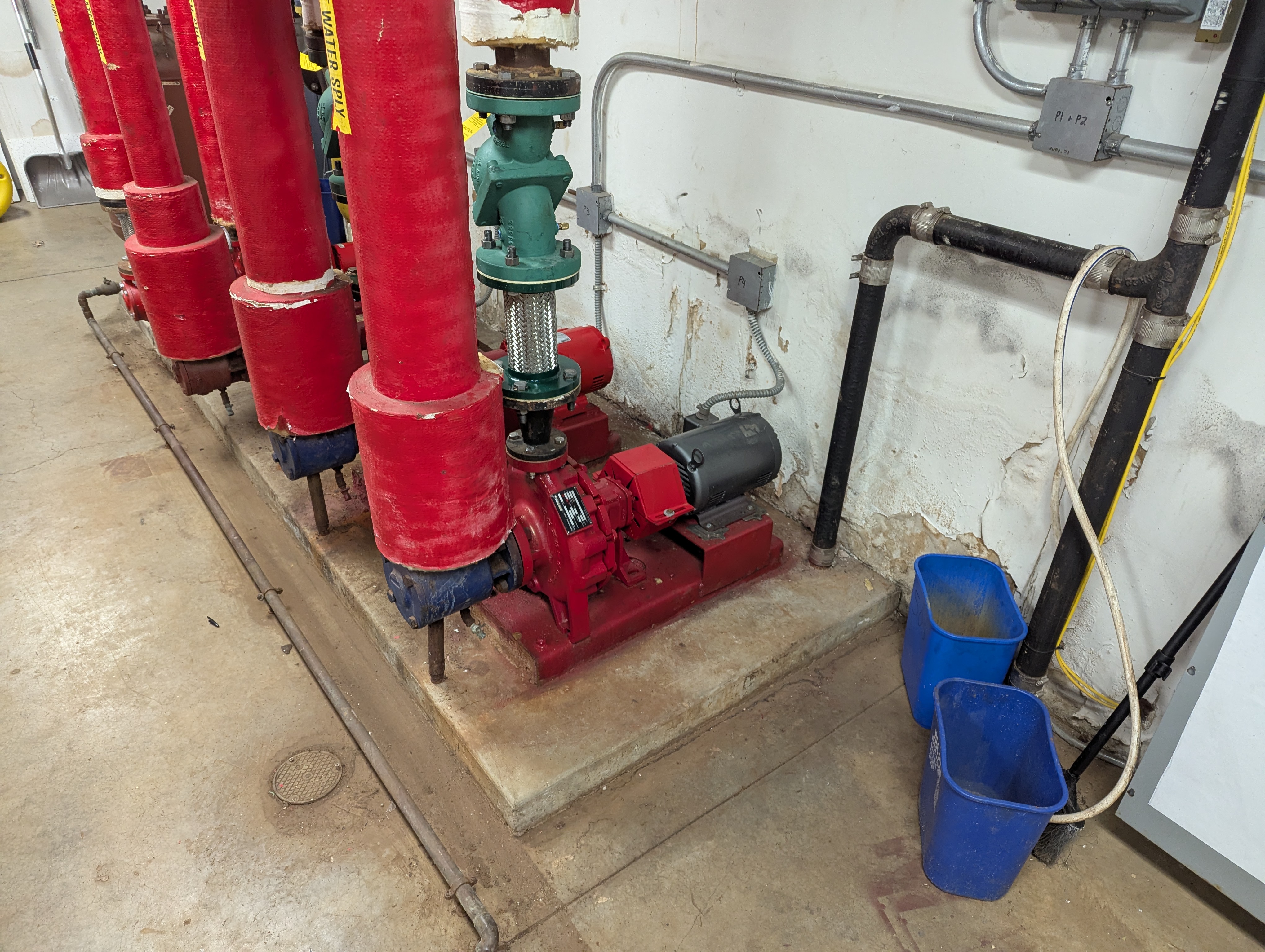 In the foreground, a red pump sits on a concrete platform next to blue buckets, while the background features industrial pipes and a white wall.