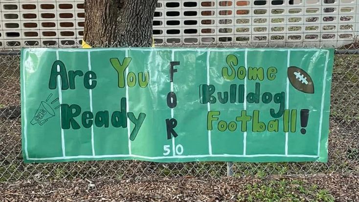 A green banner with "Are You Ready FOR Some Bulldog Football!" written in colorful letters hangs on a chain-link fence in front of a white-paneled wall and a tree.