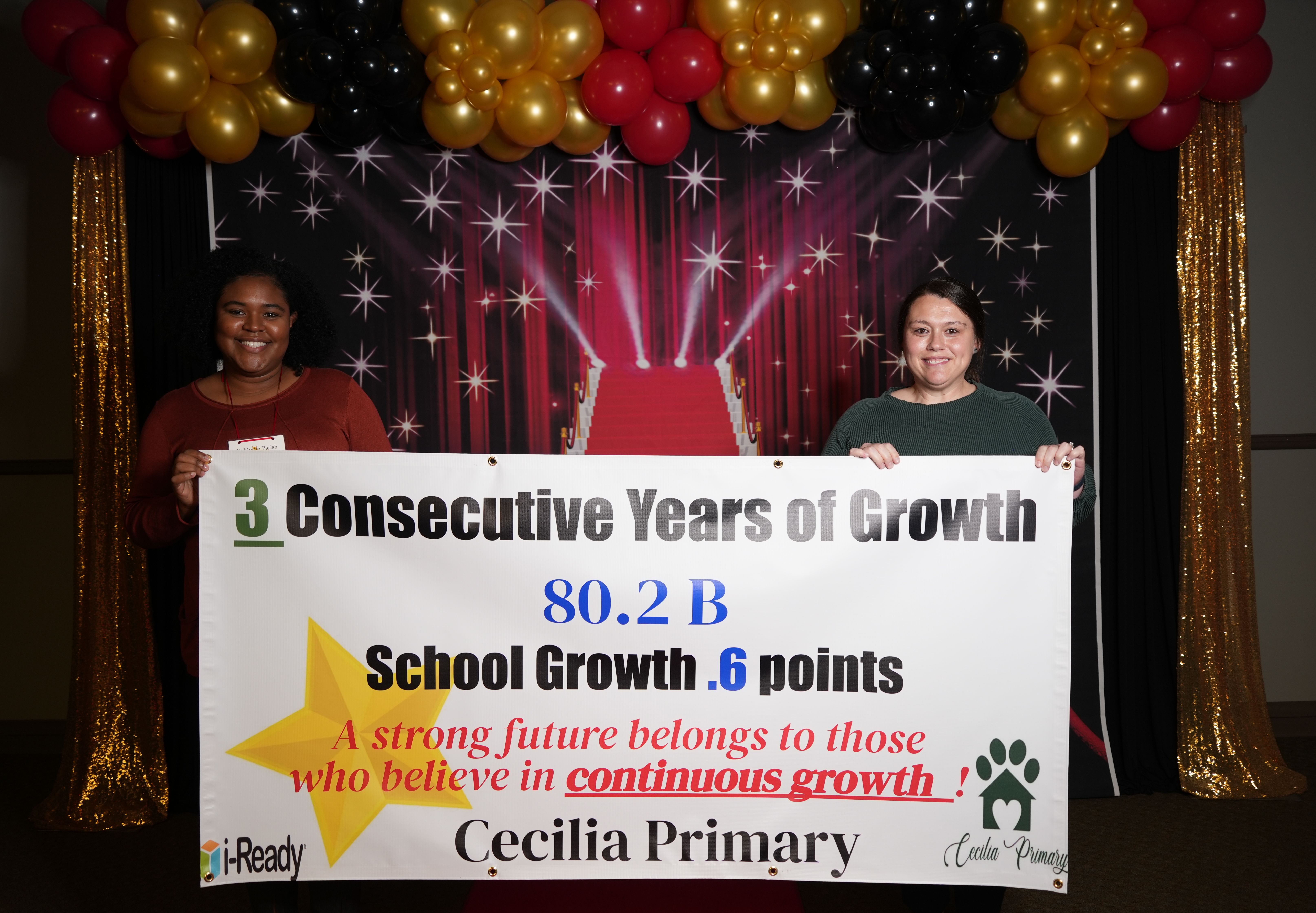 Two women stand in front of a backdrop with balloons and a red carpet, holding a banner celebrating "3 Consecutive Years of Growth" for Cecilia Primary.