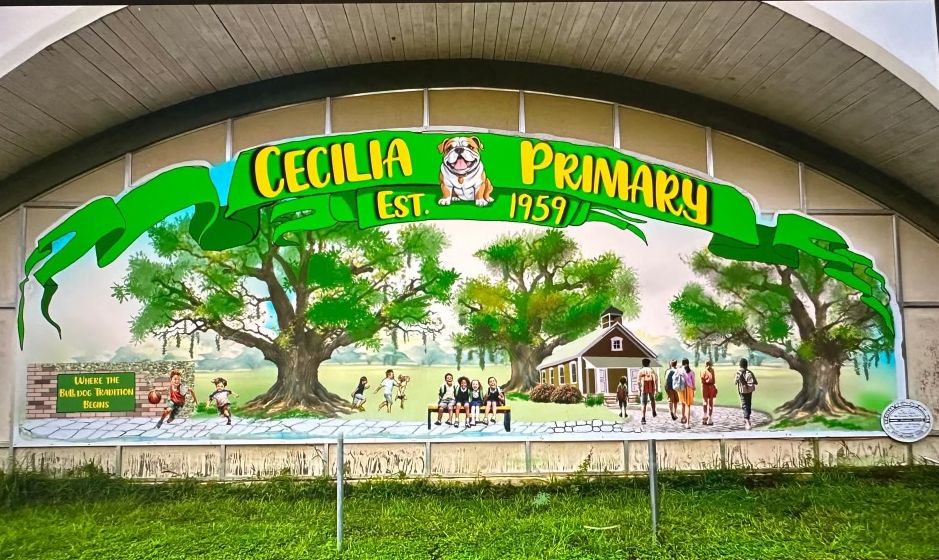 A vibrant mural on a building's exterior, featuring a banner with "CECILIA PRIMARY EST. 1959" and a bulldog, depicts children playing under large trees and a schoolhouse in the background, with green grass in the foreground.