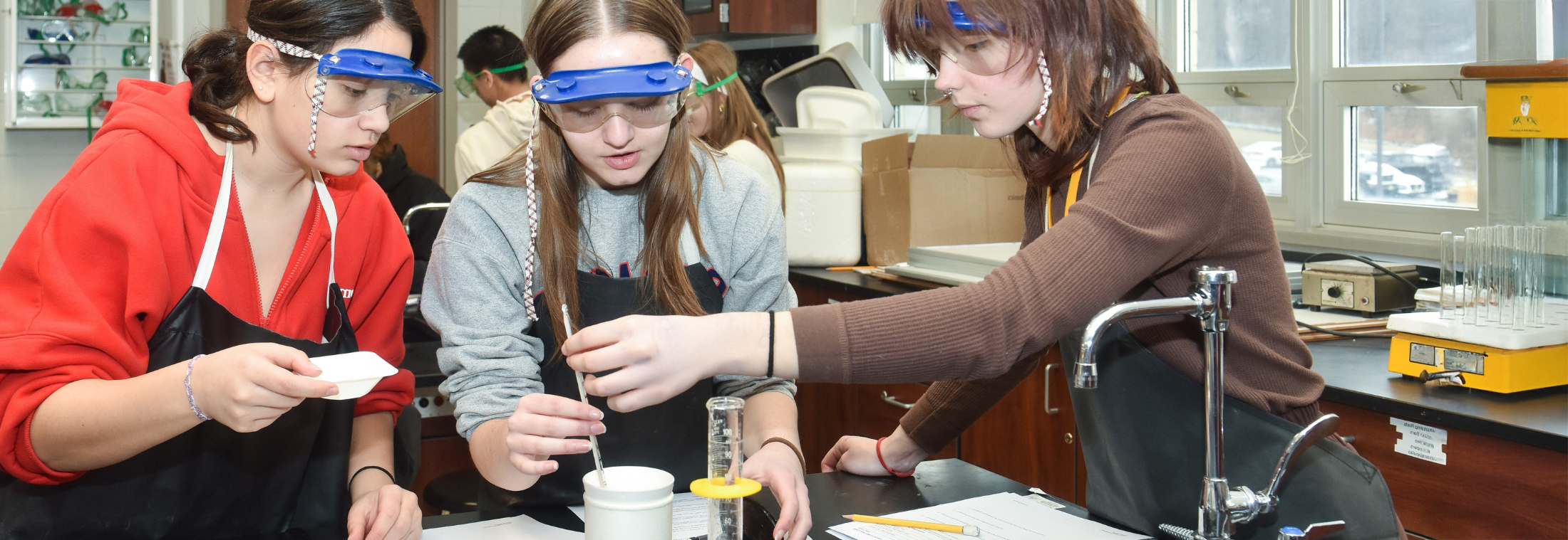 The image shows a group of young people, likely students, working together on a project or experiment in what appears to be a laboratory or classroom setting. They are wearing protective goggles and seem to be engaged in a hands-on activity involving various scientific equipment and materials.
