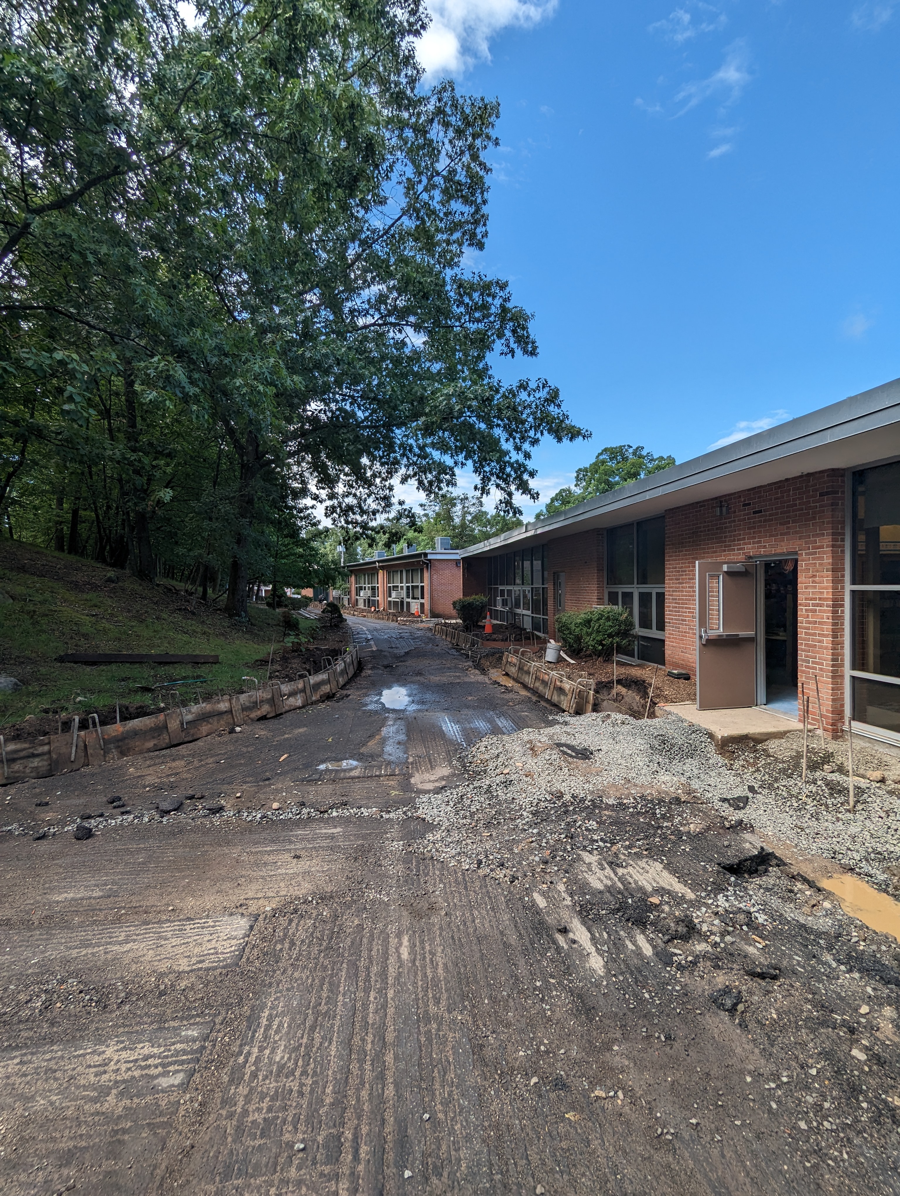 A muddy, construction-filled path leads towards a brick building with large windows, backed by a lush green hillside under a bright blue sky.