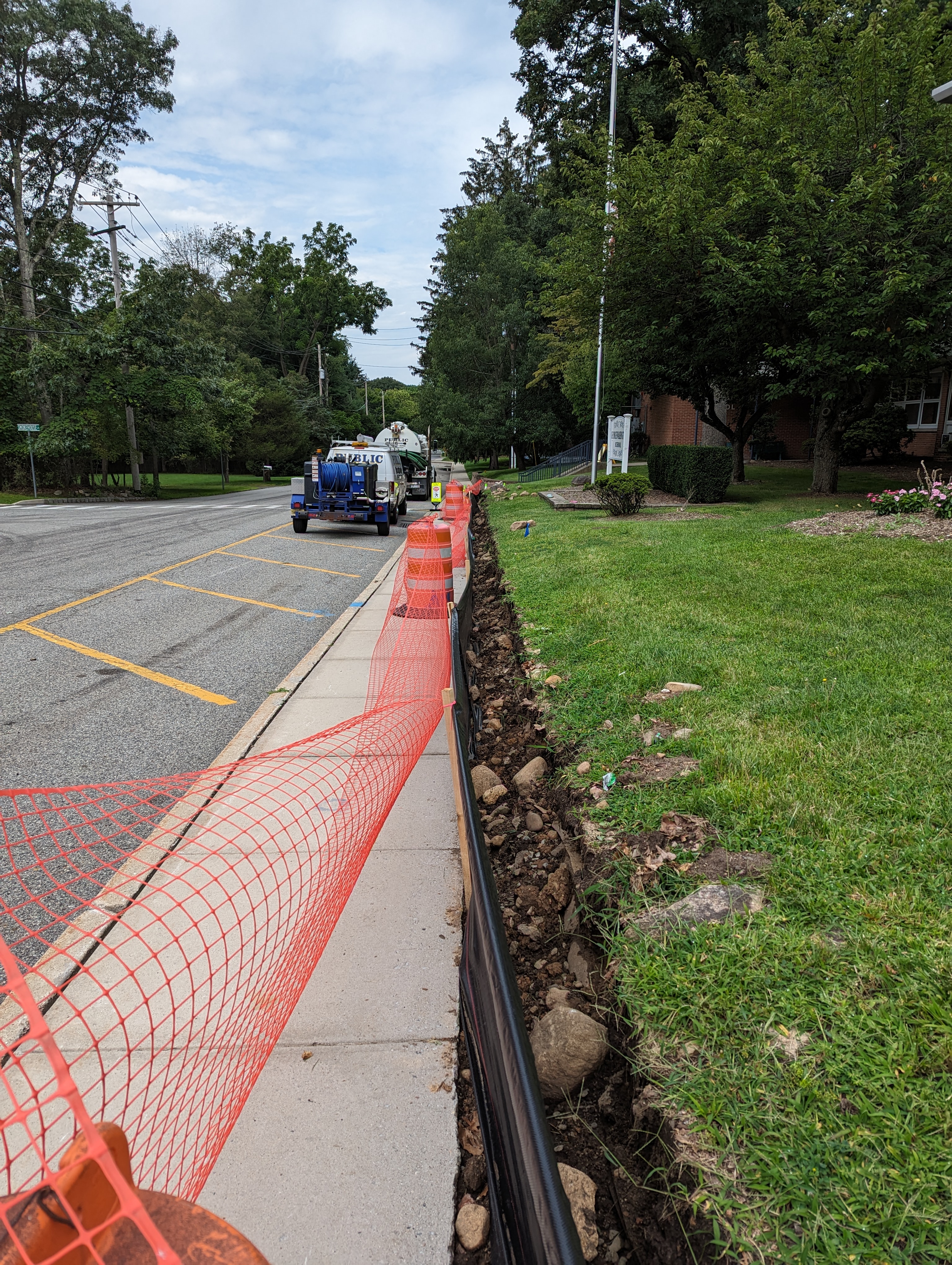 Orange safety netting and construction equipment line a sidewalk next to a freshly dug trench, with a street and trees visible in the background.