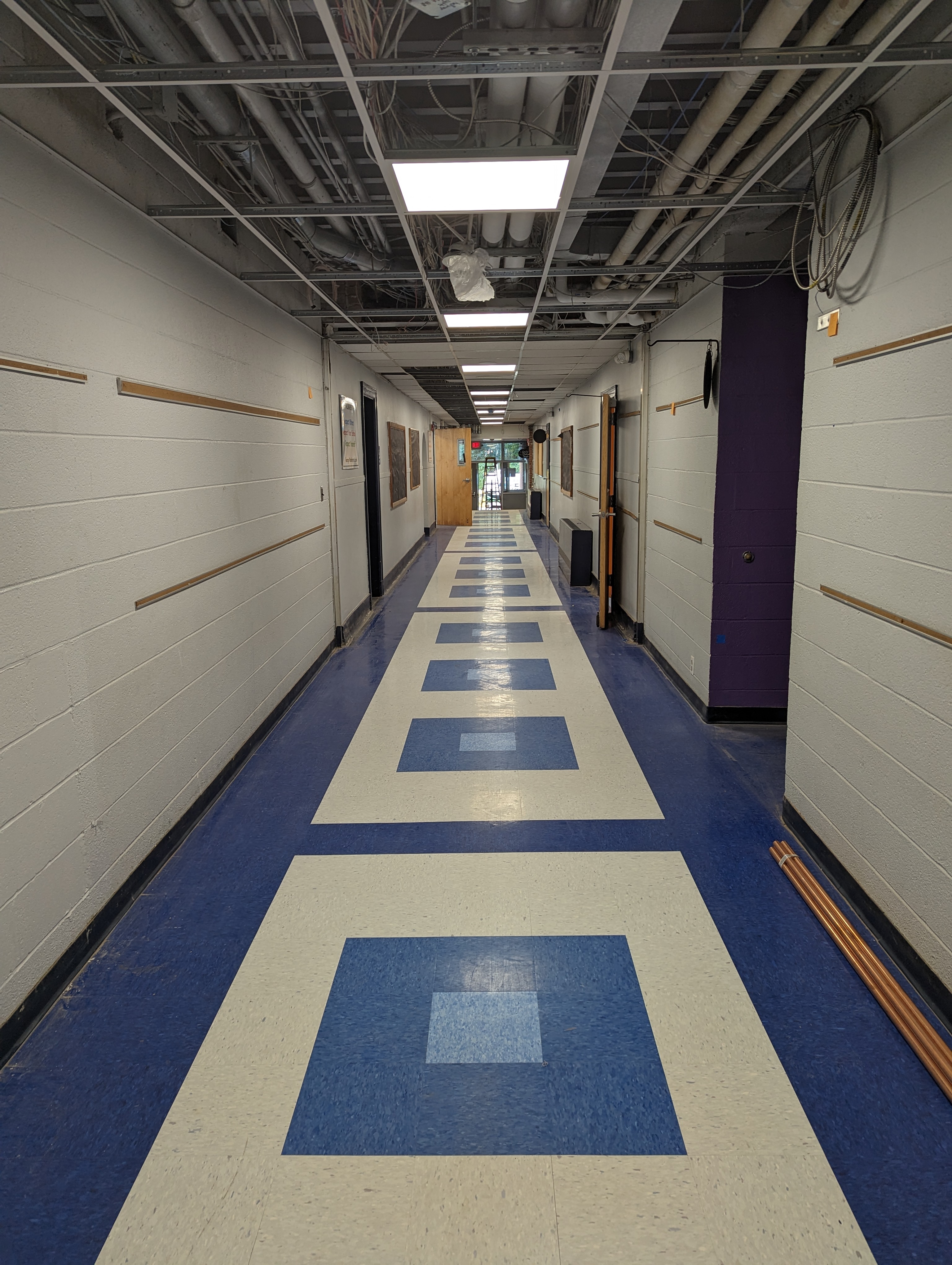 A long, brightly lit hallway with a blue and white checkered floor pattern in the foreground leads to a distant doorway, while exposed pipes and conduits crisscross the ceiling in the background.