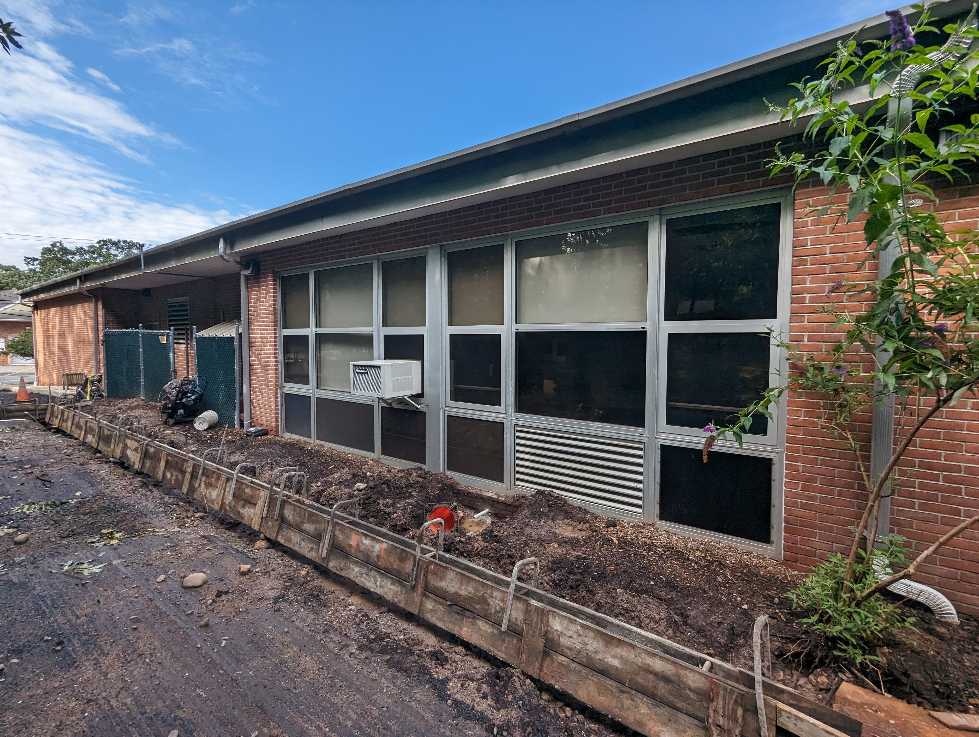 A construction site with a brick building and large windows in the background is in the foreground, with a wooden barrier and dirt.