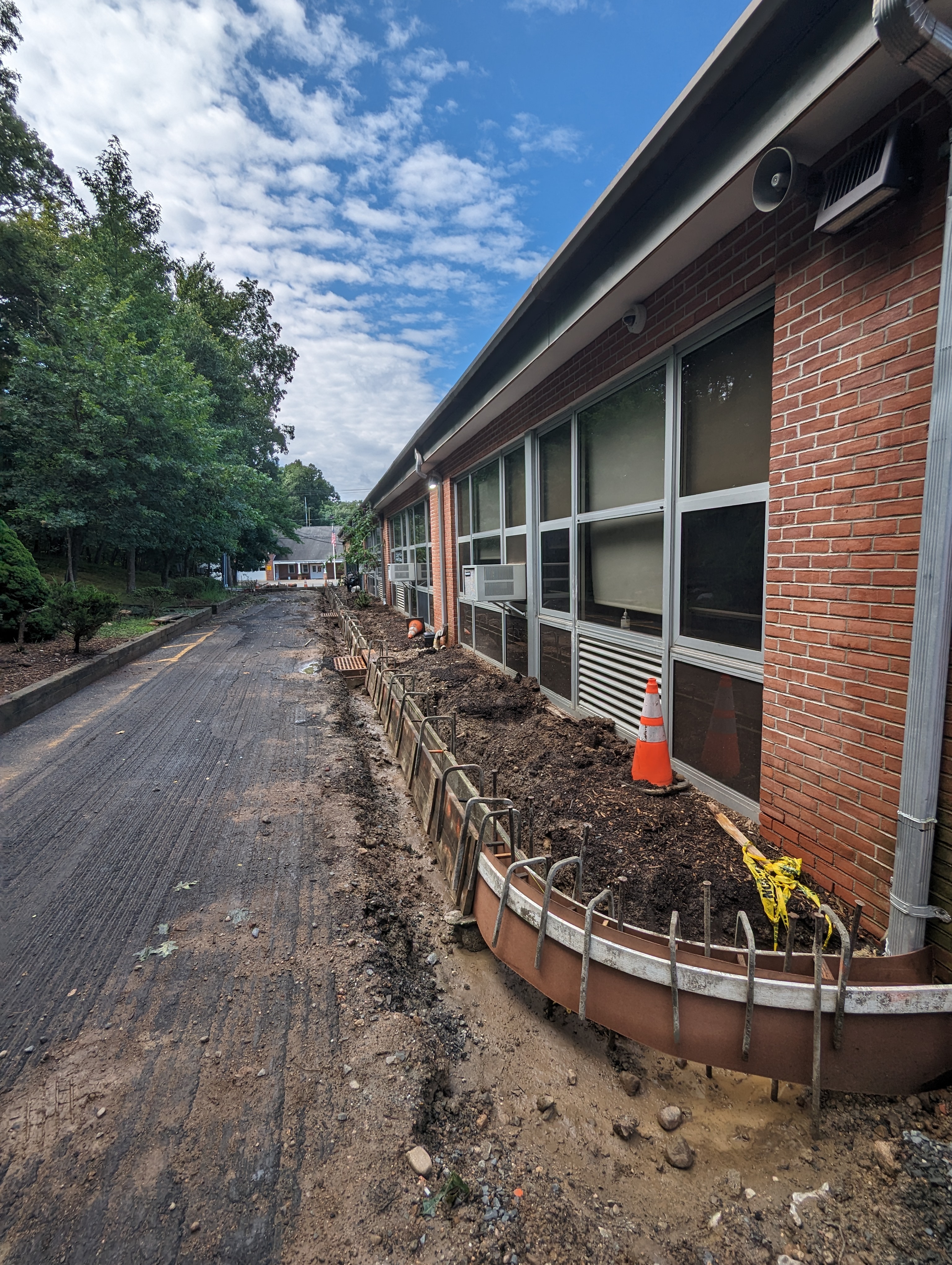 Construction work is underway along the side of a brick building with a paved walkway in the foreground and trees in the background.
