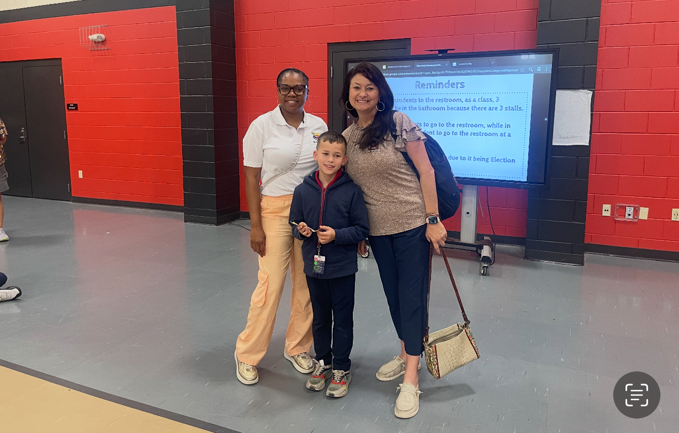 Two women and a boy stand in a gymnasium with a large screen displaying reminders in the background.